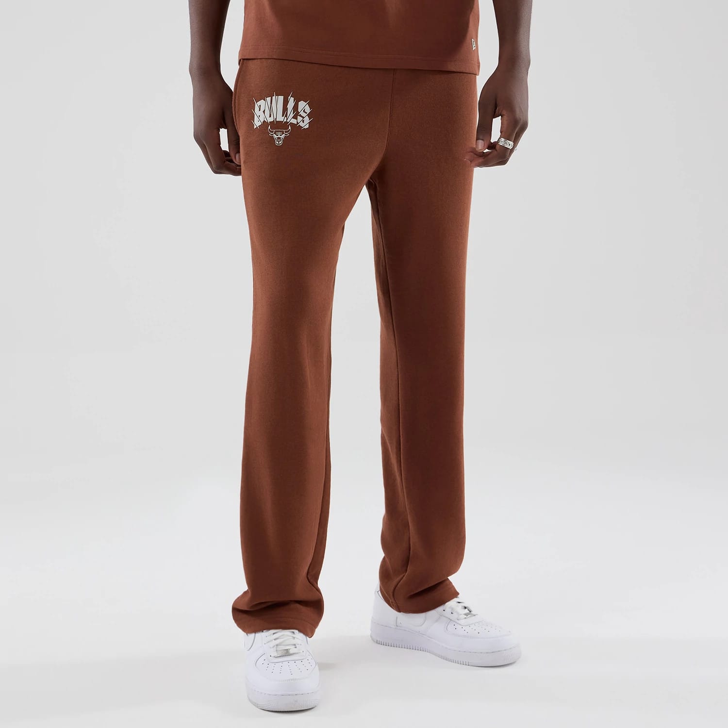 New Era Chicago Bulls NBA Core Plus Brown Straight Leg Joggers, front view on a model showing the Bulls team name and logo in white on the upper left leg, with a relaxed straight leg cut and paired with white trainers.