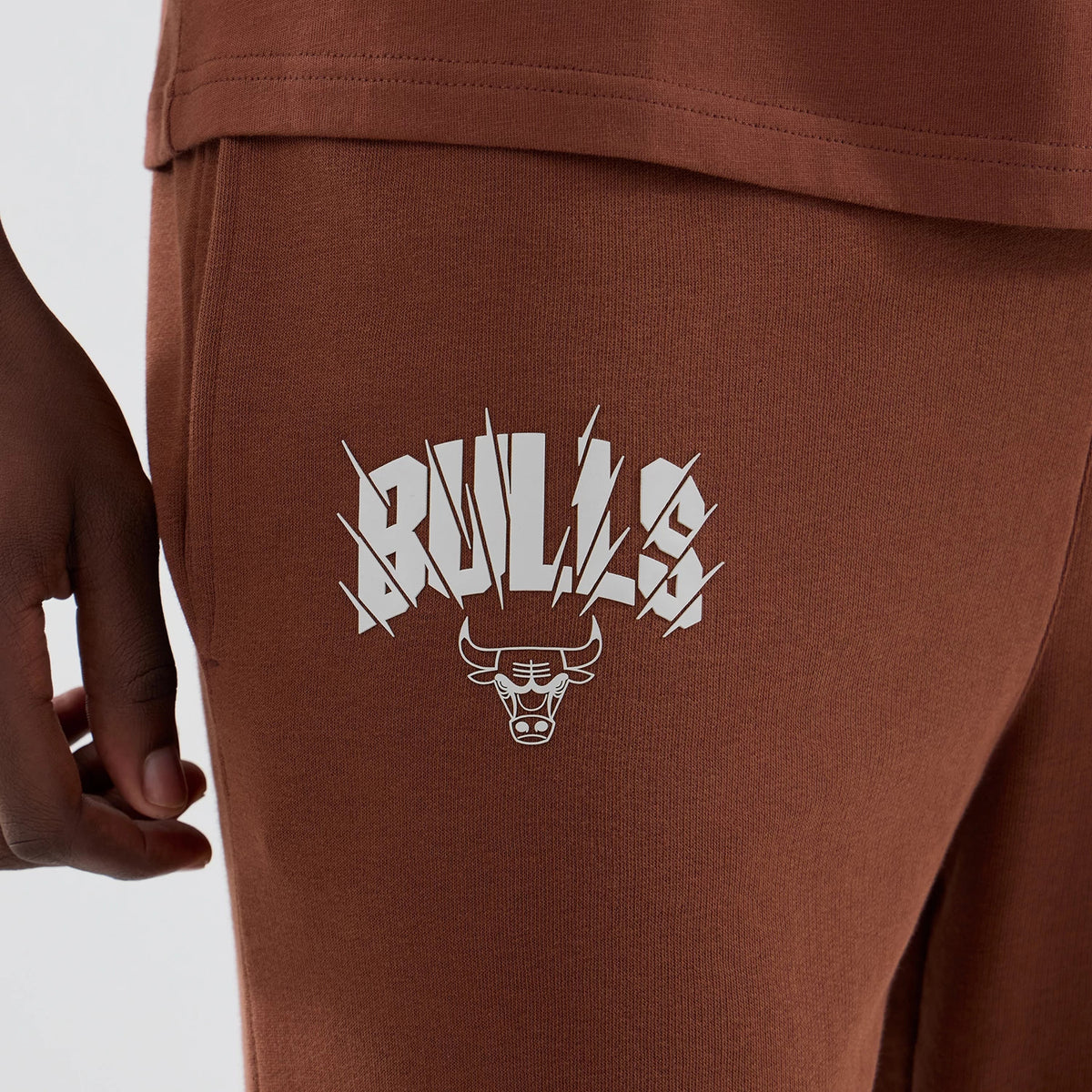 New Era Chicago Bulls NBA Core Plus Brown Straight Leg Joggers, side view showing the white Bulls logo with distinctive lettering and team mascot printed on the left thigh, in a rich brown fabric with a straight-leg cut.