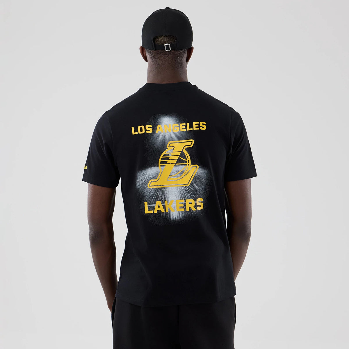 New Era LA Lakers NBA Court Graphic Black T-Shirt, back view on a model featuring prominent yellow LOS ANGELES LAKERS text above and below a large graphic L over a faded white graphic, small embroidered logo on the sleeve and a black cap.