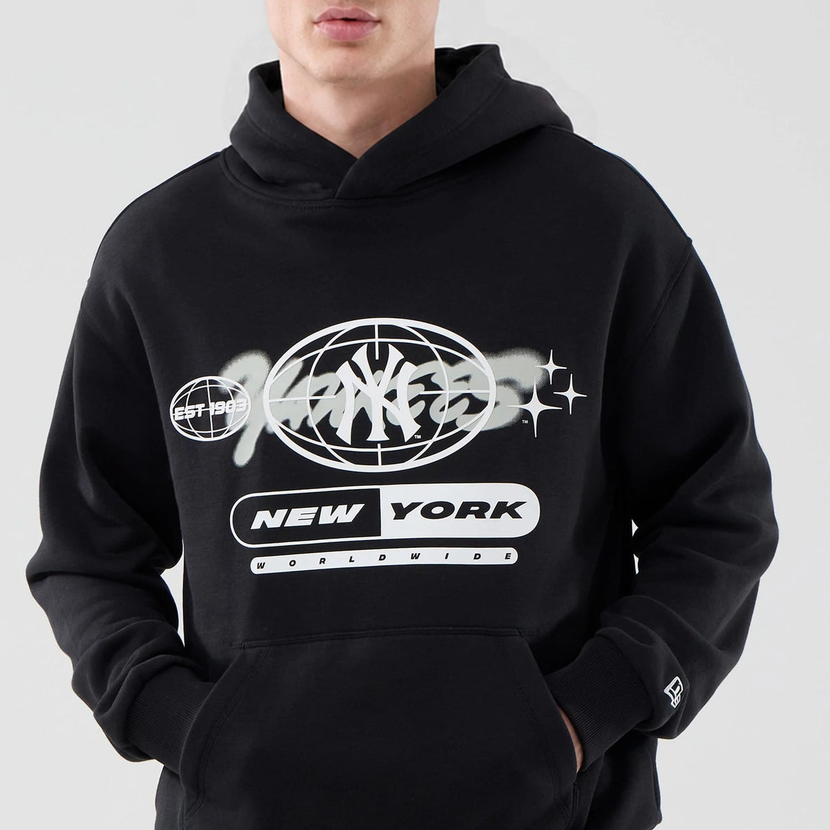 New Era New York Yankees MLB Logo Graphic Black Oversized Pullover Hoodie, front view showing the white graphic with NY Yankees symbol and NEW YORK text on the chest, a kangaroo pocket, and an embroidered New Era logo on the left sleeve cuff.
