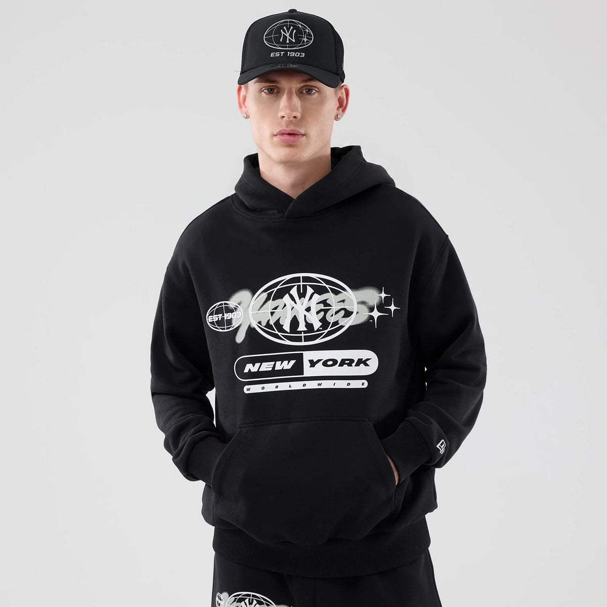 New Era New York Yankees MLB Logo Graphic Black Oversized Pullover Hoodie, front view on-model featuring a large central graphic with Yankees and New York text, abstract globe design, and a small embroidered New Era logo on the left sleeve cuff.