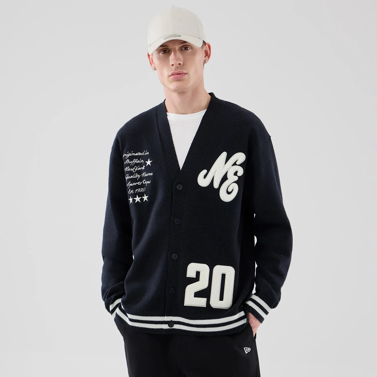 New Era Heritage Navy Cardigan, front view on a model, featuring large embroidered white lettering NE on the left chest, script and stars on the right chest, and the number 20 on the lower right side above ribbed striped cuffs.