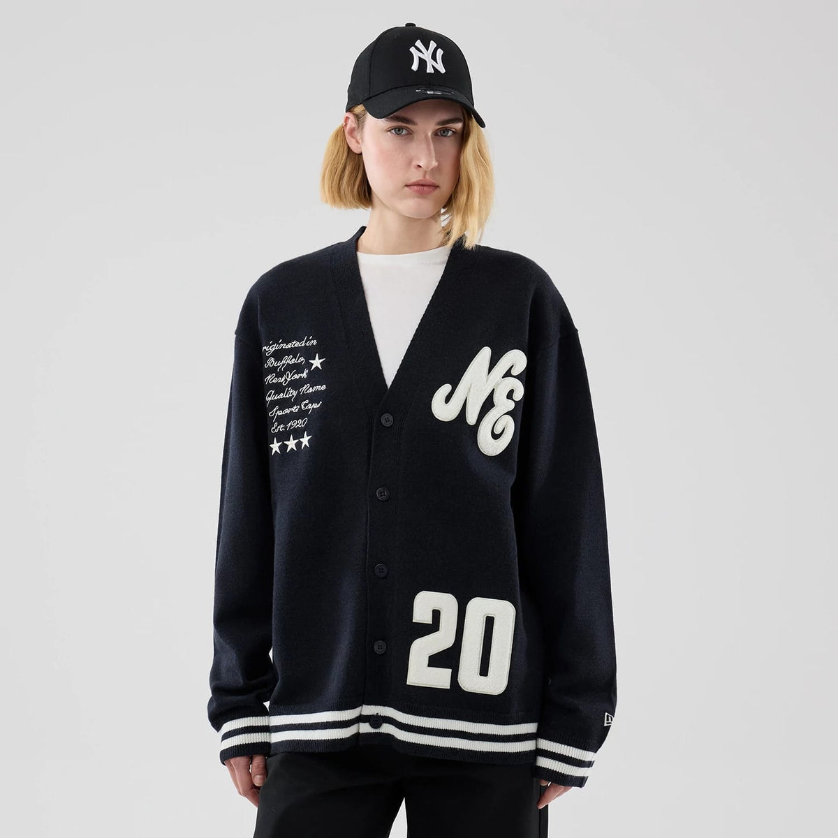 New Era Heritage Navy Cardigan, front view on a model with a black cap, showing large white NE lettering on the left chest, intricate white script and star embroidery on the right side, and 20 numbering along the bottom hem.