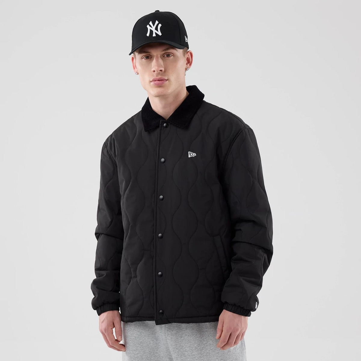 New Era Quilted Coaches Black Jacket, front view on a model featuring a quilted pattern with button closure, a small embroidered New Era logo on the left chest, and a black collar, styled with a black cap and grey trousers.