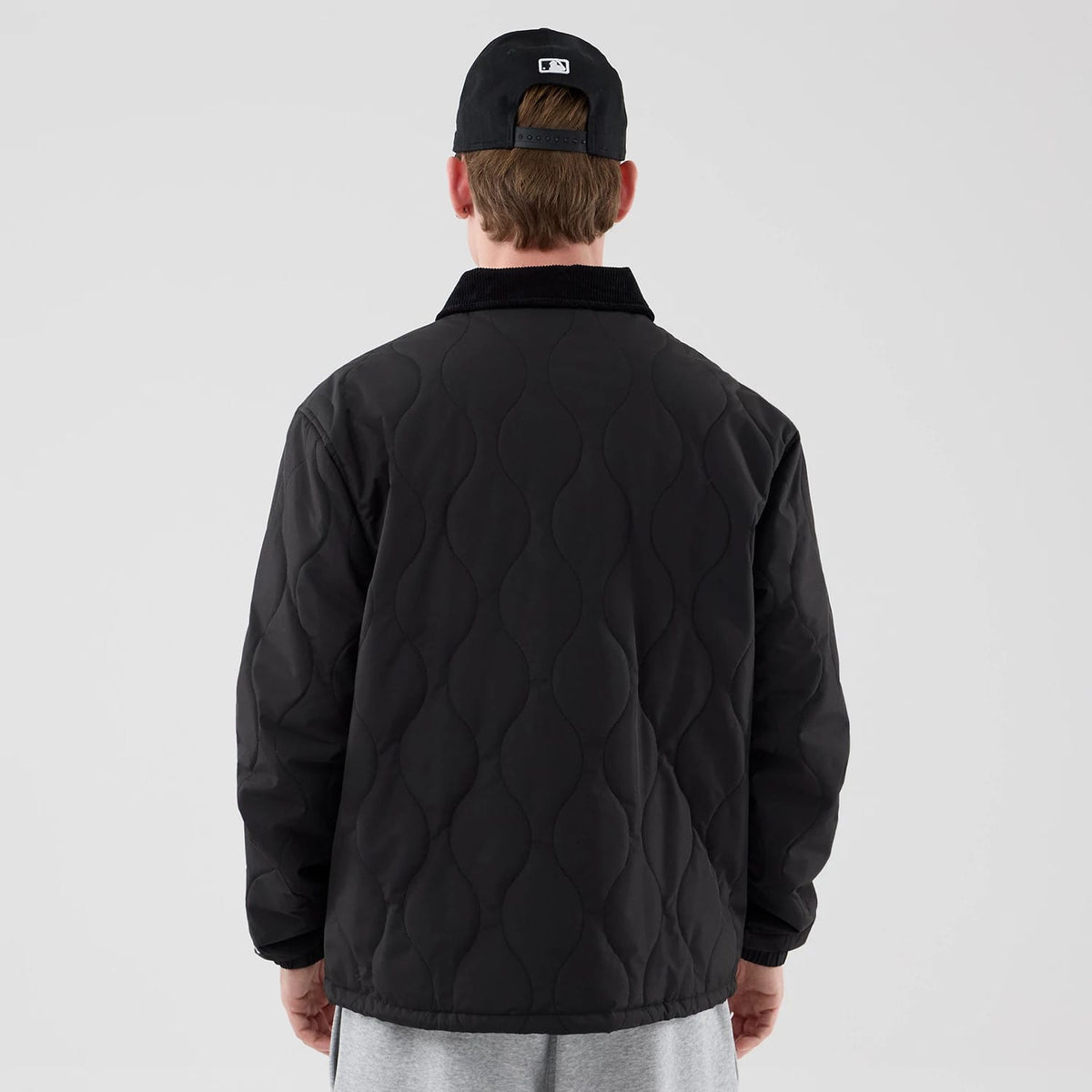 New Era Quilted Coaches Black Jacket, back view on a model showing the distinctive wavy quilted pattern across the back, a solid black collar, and pairing with a black cap worn backwards featuring an embroidered logo above the closure.