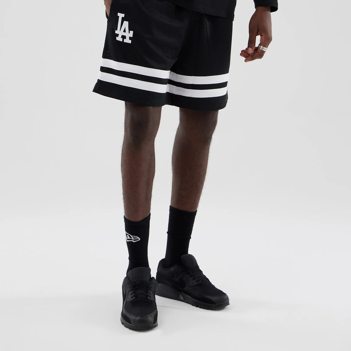 New Era LA Dodgers MLB Mesh Black Oversized Shorts, front view on a model with black trainers and socks, featuring a white embroidered LA logo on the left thigh, and two horizontal white stripes across the lower part of the shorts.