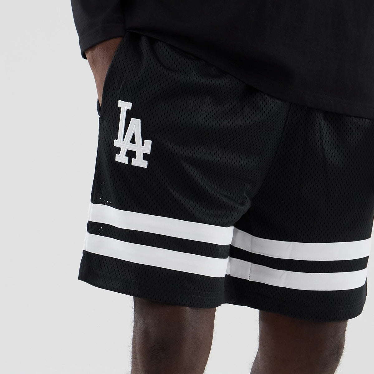 New Era LA Dodgers MLB Mesh Black Oversized Shorts, front view showing the white embroidered LA logo on the left leg, two white horizontal stripes near the hem, and a relaxed fit, styled with a simple black top.