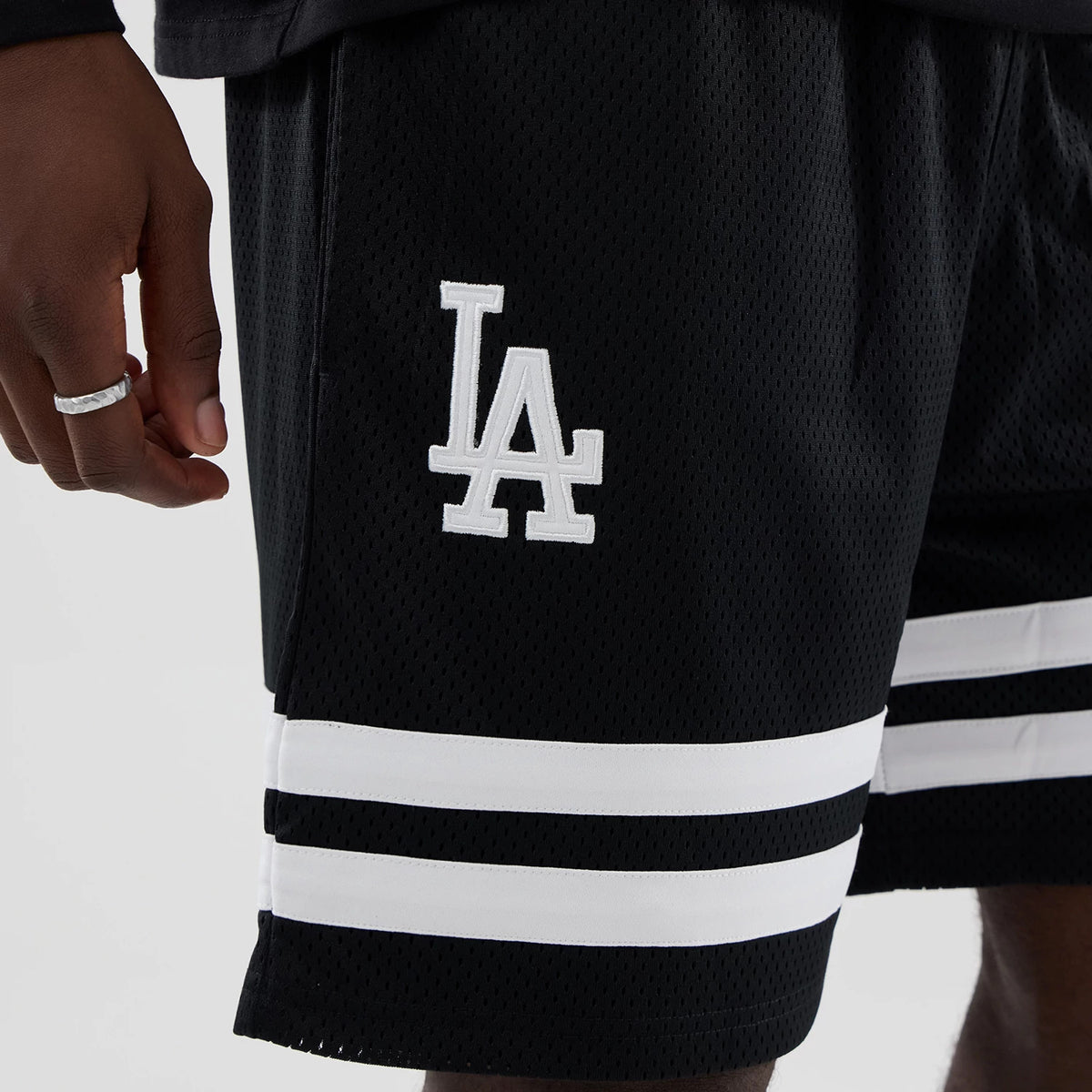 New Era LA Dodgers MLB Mesh Black Oversized Shorts, front view showing the prominent white embroidered LA logo on the side, two horizontal white stripes near the hem, and designed with a mesh texture for added style.