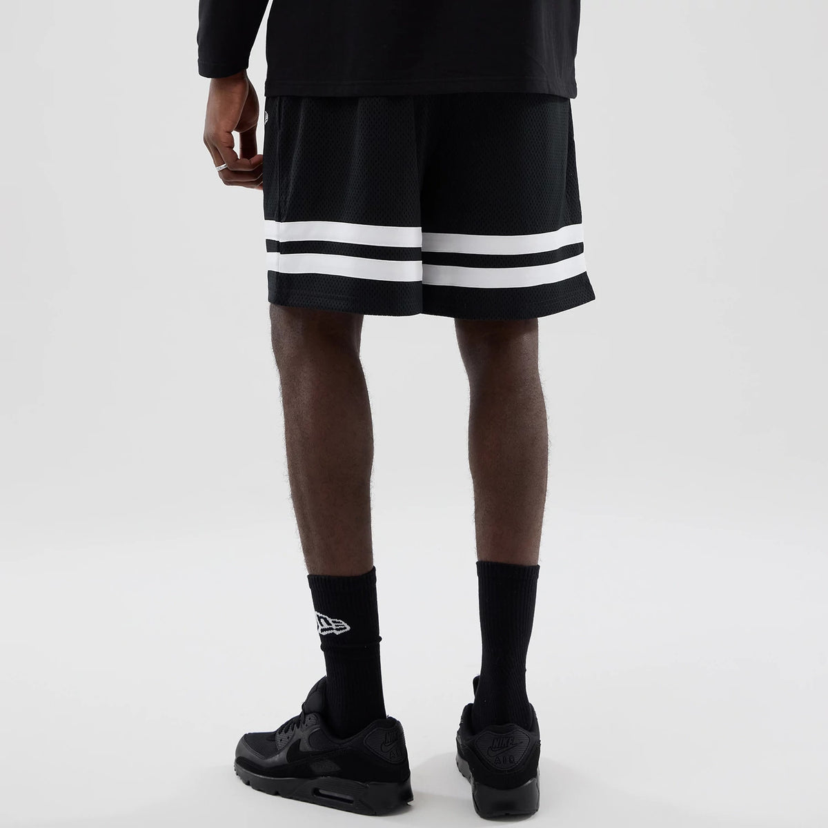 New Era LA Dodgers MLB Mesh Black Oversized Shorts, back view showing two white horizontal stripes near the hem on a model wearing black trainers and socks, highlighting the relaxed fit and distinctive design of the shorts.
