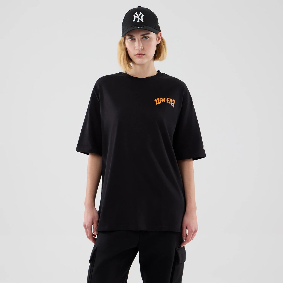 New Era Hot Dog Graphic Black Oversized T-Shirt, front view on a model paired with a black cap, featuring orange New Era lettering on the left chest, oversized fit with short sleeves, and sleeve detail.
