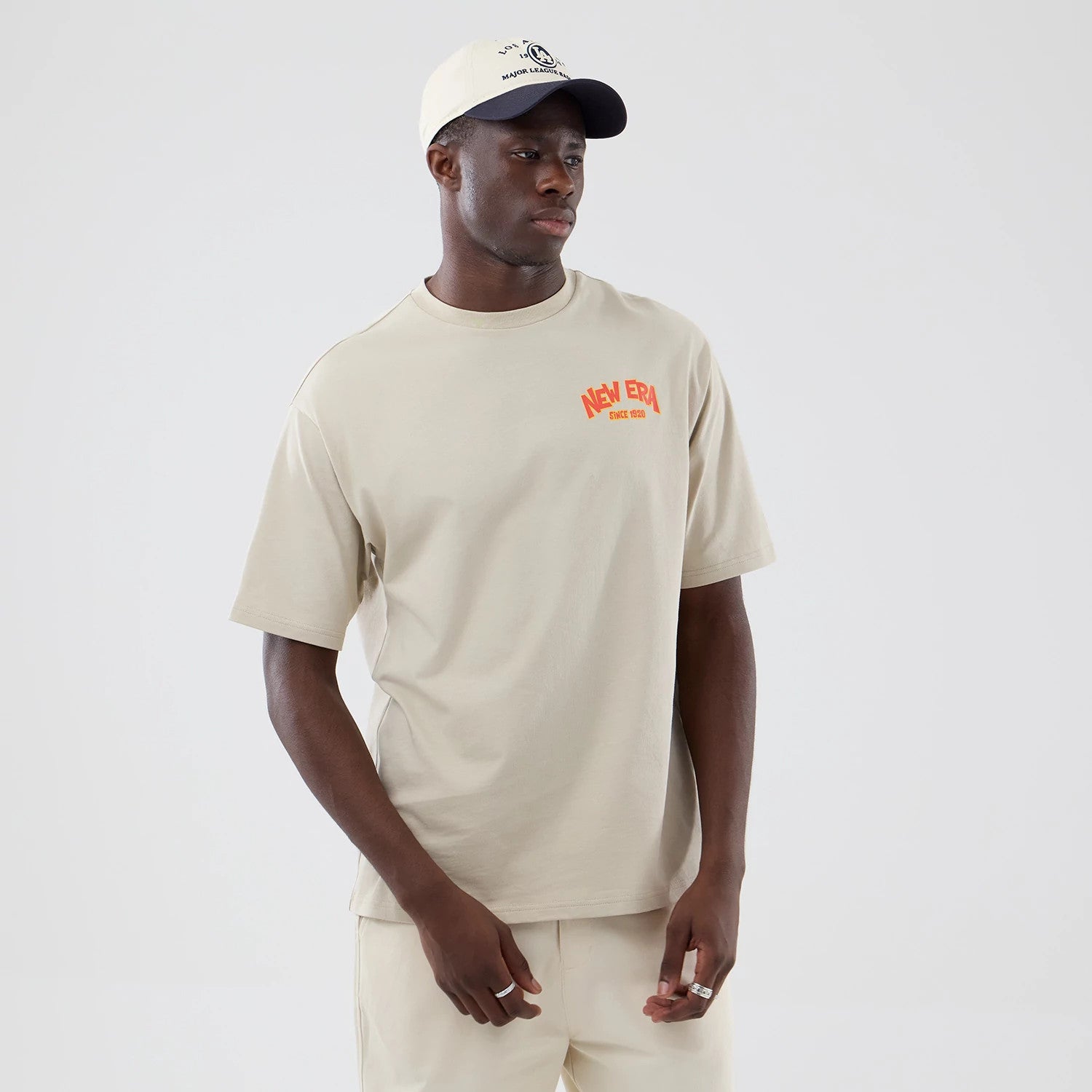 New Era Food Graphic Light Beige Oversized T-Shirt, front view on a model styled with a contrasting cream cap, featuring an orange printed New Era logo on the left chest, short sleeves, and a relaxed fit with plain light beige fabric.
