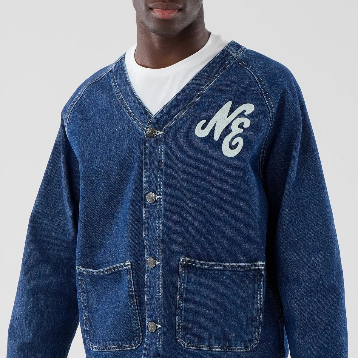 New Era Denim Dark Blue Worker Jacket, front view on-model showing a deep blue denim with a button closure, an embroidered NE logo on the chest, and two large front pockets, styled over a plain white T-shirt.