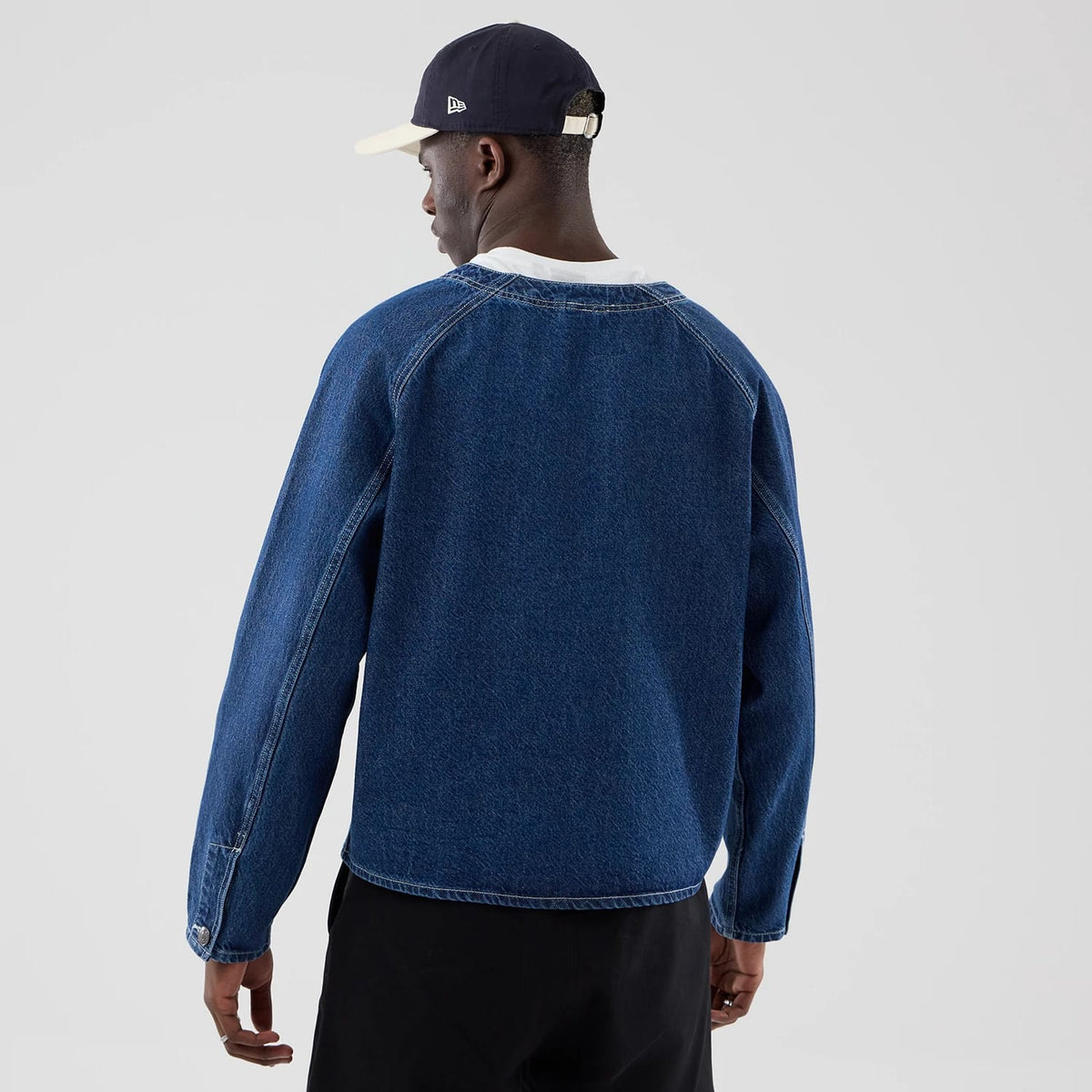 New Era Denim Dark Blue Worker Jacket, back view on a model styled with a navy and white cap, showcasing the simple dark blue denim design with stitching along the raglan sleeves and hem, and button details at the cuffs.