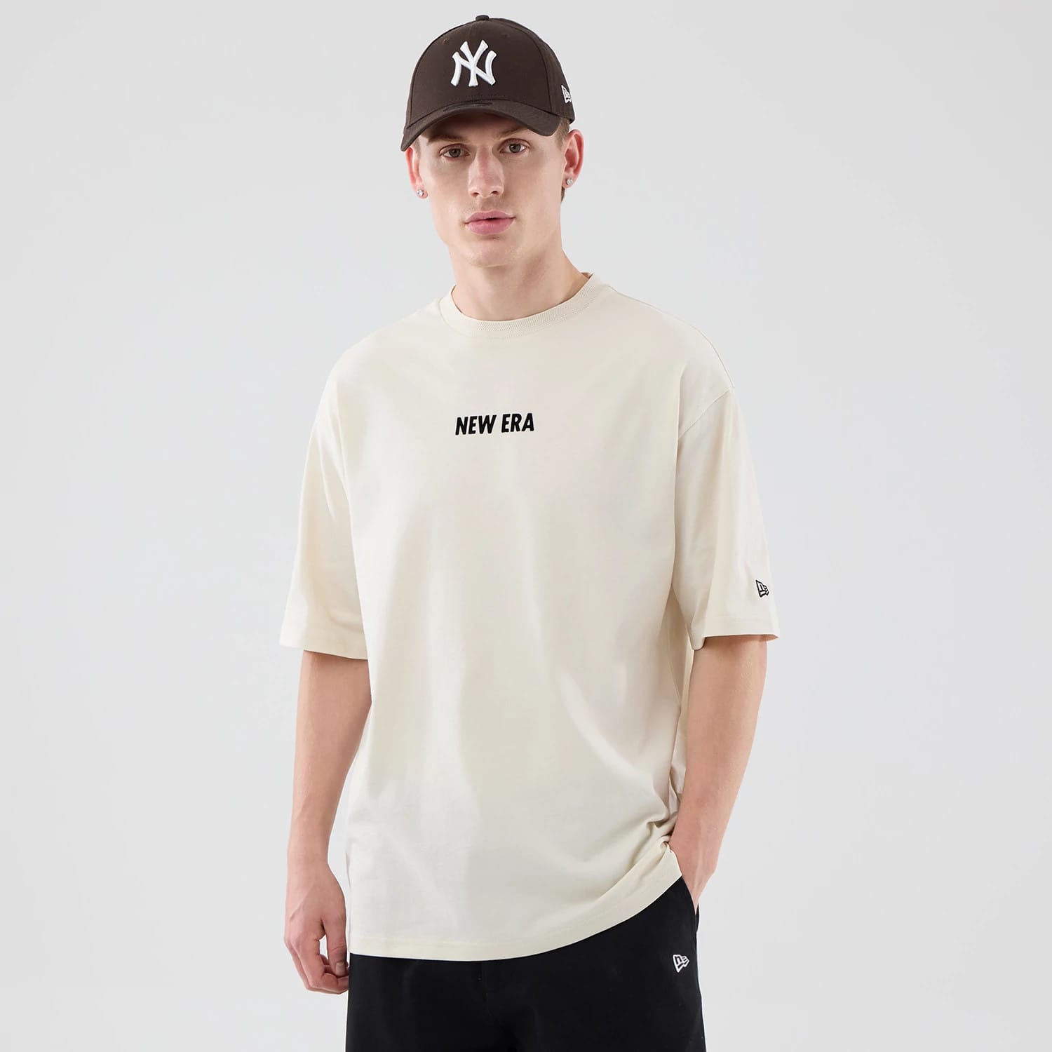 New Era Cap Graphic Cream Oversized T-Shirt, front view on-model showing the cream T-shirt with black embroidered NEW ERA text centred on the chest, a New Era logo on the left sleeve and paired with black trousers featuring a matching logo.