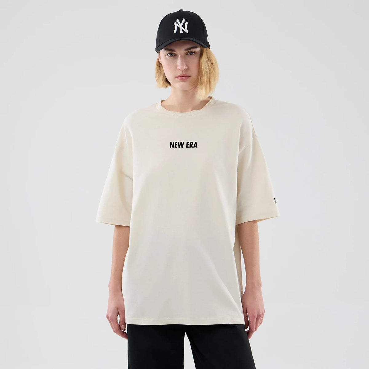 New Era Cap Graphic Cream Oversized T-Shirt, front view on-model showing a relaxed fit with the words NEW ERA printed in black across the chest, short sleeves reaching the elbows, and a small embroidered New Era logo on the left sleeve.