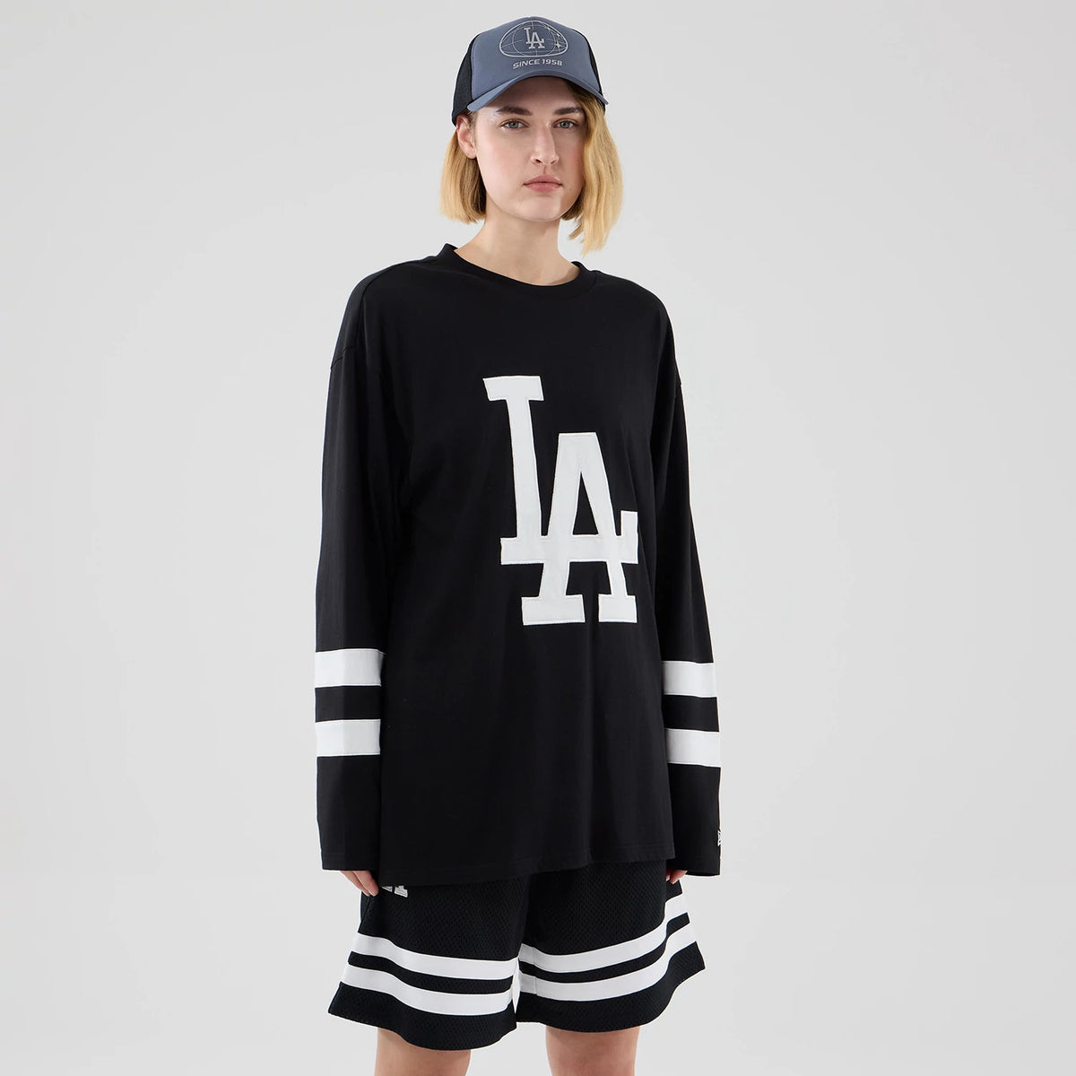 New Era LA Dodgers MLB Black Oversized Long Sleeve T-Shirt, front view on a model styled with matching shorts and a cap, prominently featuring a large white printed LA logo on the chest and horizontal white stripes on the sleeves and shorts hem.