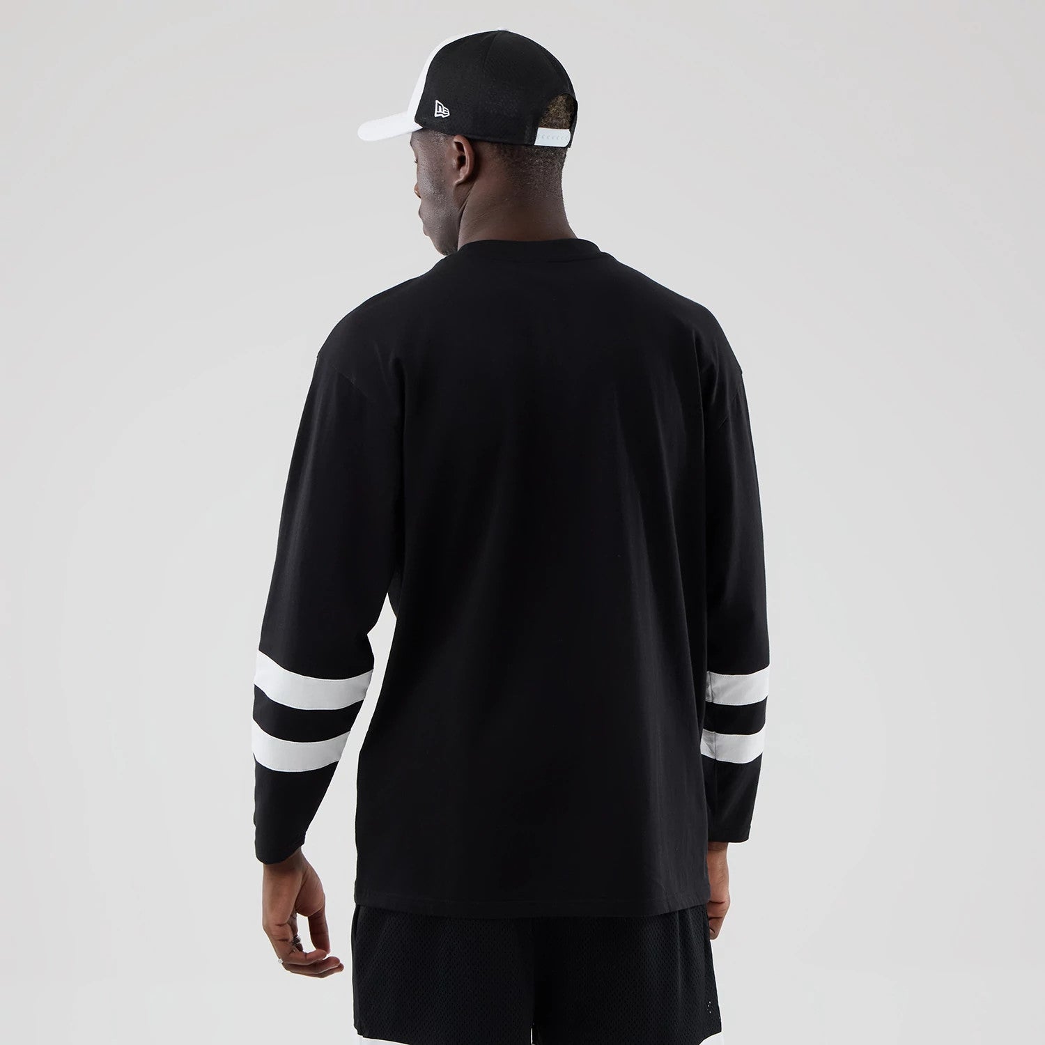 New Era LA Dodgers MLB Black Oversized Long Sleeve T-Shirt, back view on a model showcasing a simple black design with distinctive white striped on the lower sleeves, paired with matching black shorts and a black and white cap.