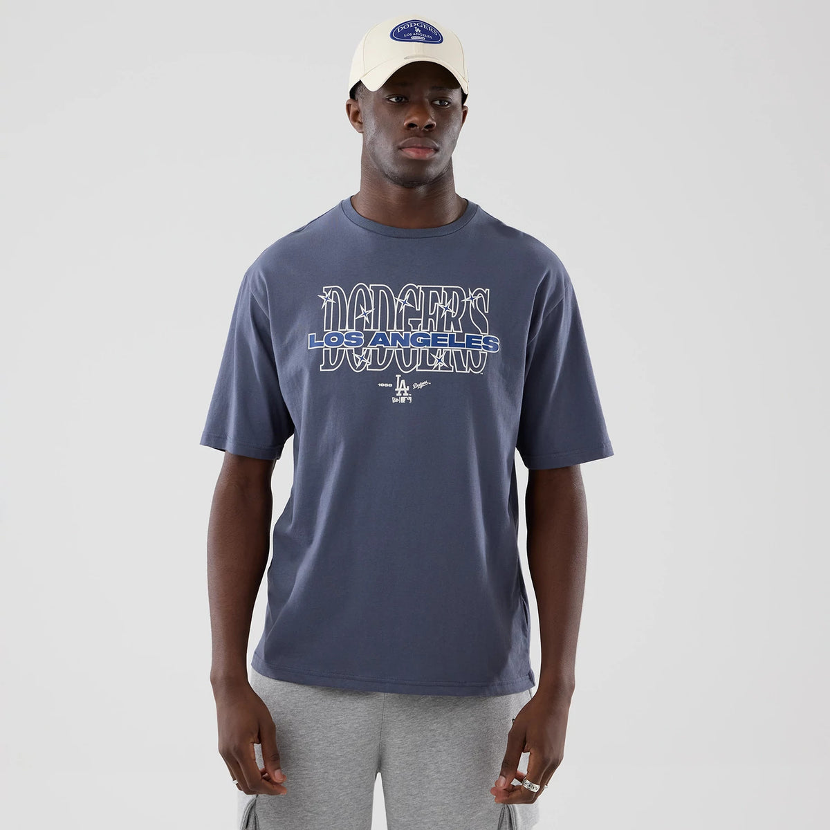 New Era LA Dodgers MLB Graphic Grey Oversized T-Shirt, front view on a model showcasing a Dodgers Los Angeles graphic in blue and white across the chest, complemented by small logo details beneath, styled with a cap and grey shorts.