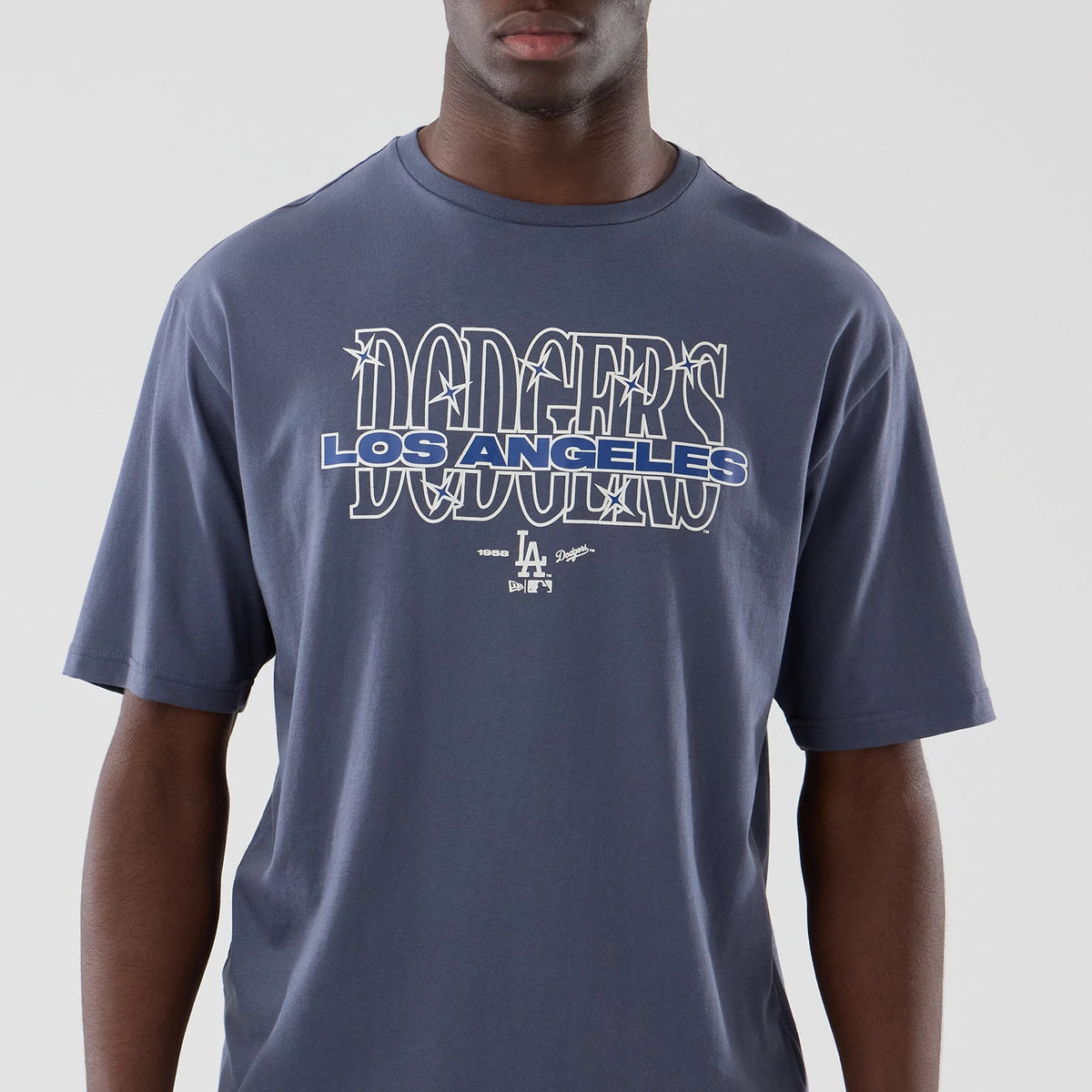 New Era LA Dodgers MLB Graphic Grey Oversized T-Shirt, front view on-model showing the Dodgers lettering in white outlined on the chest, with a blue Los Angeles imprint underneath and a small printed LA team logo at the bottom centre.