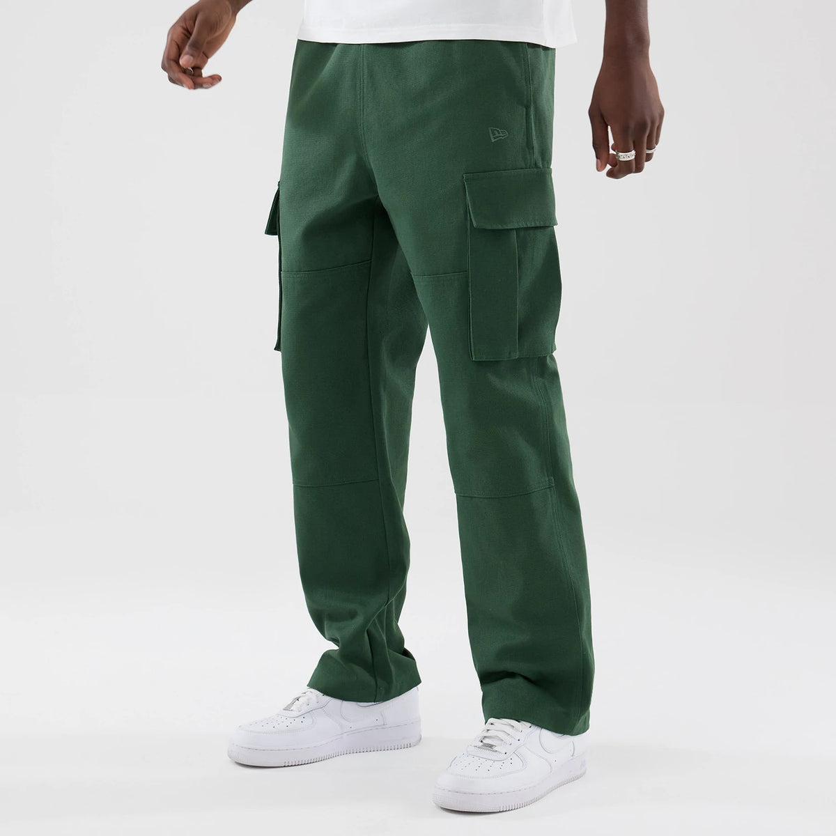 New Era Dark Green Cargo Trousers, front view on a model showing two large flap pockets on each leg, New Era logo embroidered on the right hip, and loose-fitting legs extending down to white trainers.