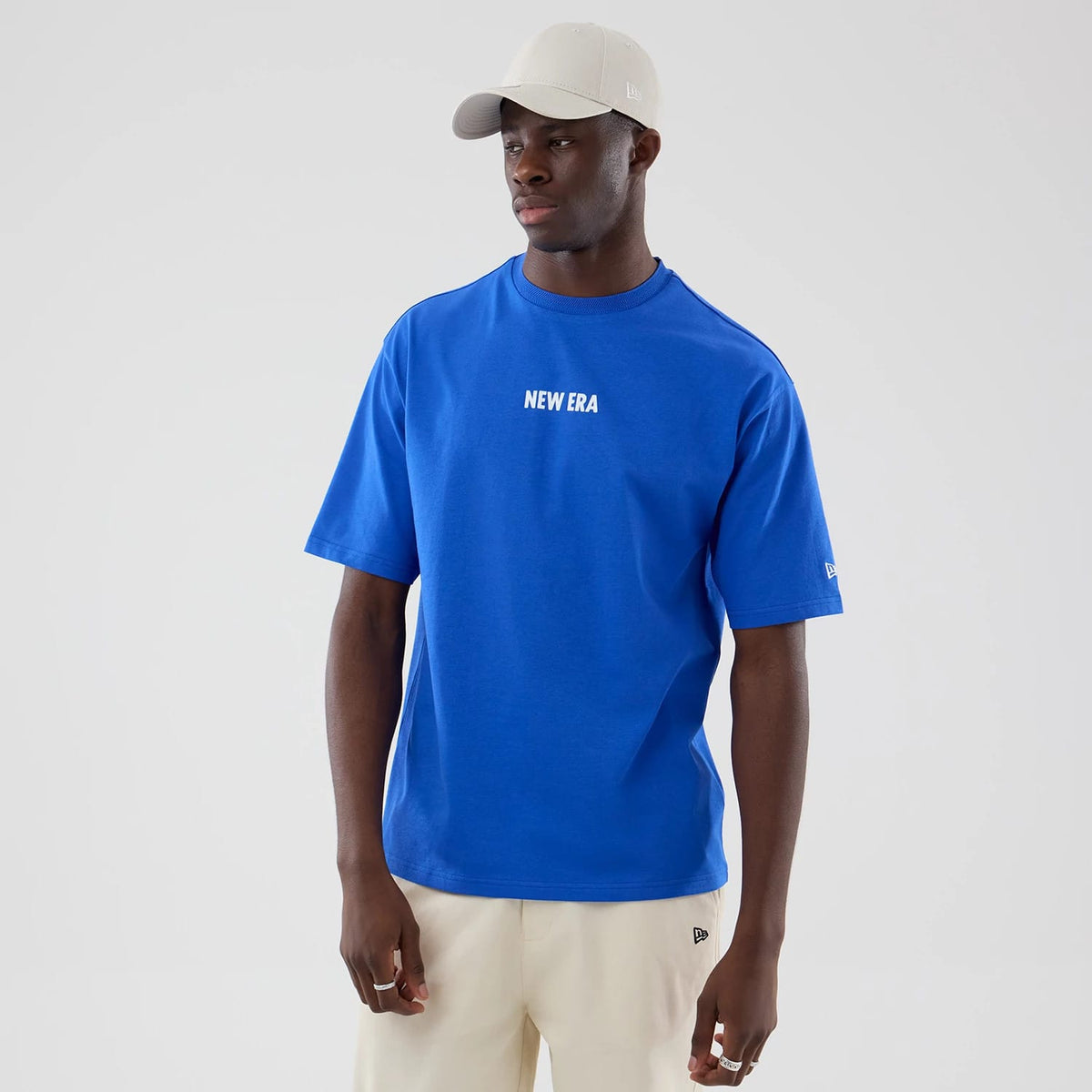 New Era Cap Graphic Bright Blue Oversized T-Shirt, front view on a model wearing a light-coloured cap, featuring a white embroidered New Era logo centred on the chest and another logo embroidered on the left sleeve above the elbow.