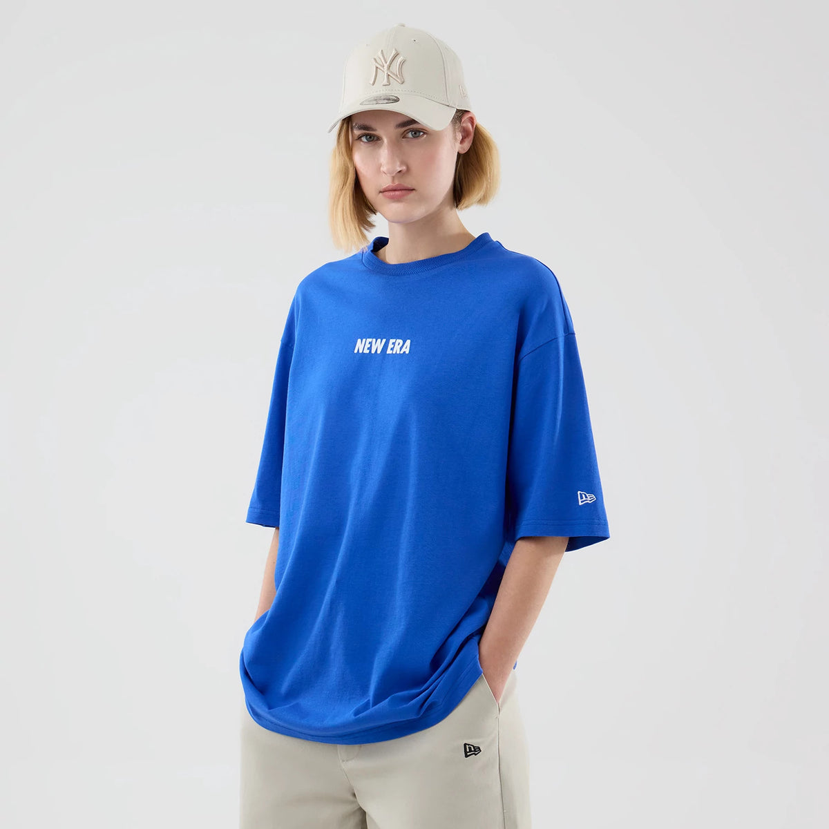 New Era Cap Graphic Bright Blue Oversized T-Shirt, front view on-model showing a blue design with the white printed New Era logo centred on the chest, the New Era embroidered logo on the left sleeve, styled with a light cap and grey trousers.