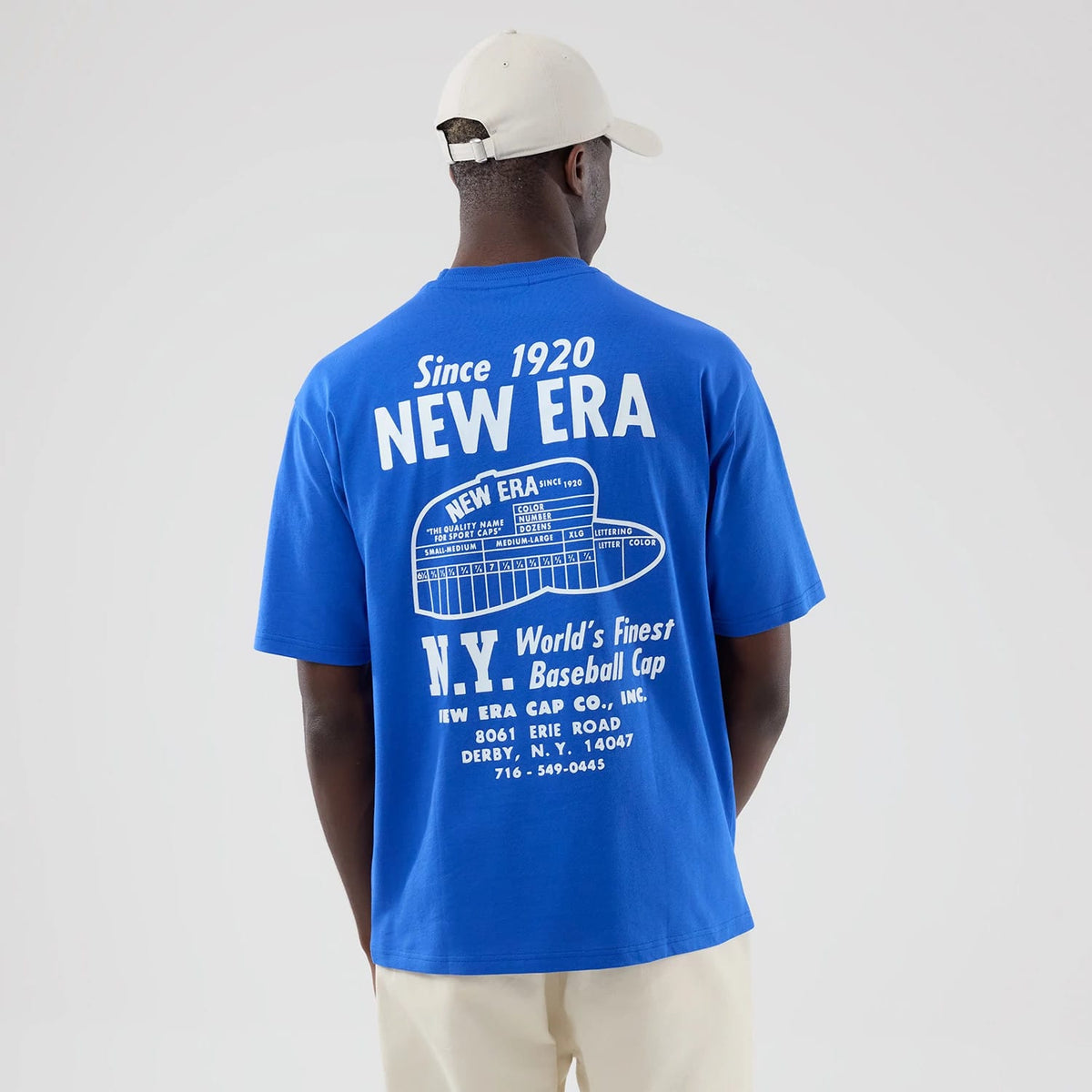 New Era Cap Graphic Bright Blue Oversized T-Shirt, back view showing white retro text and illustrative graphics displaying New Era branding, with Since 1920 and other nostalgic elements, worn by a model pairing it with a neutral cap.