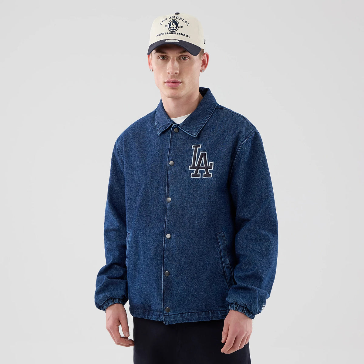 New Era LA Dodgers MLB Coaches Dark Blue Jacket, front view on-model showing a dark blue jacket with a classic collar, silver button-up front, and an embroidered white LA logo on the left chest, styled with a cream cap and dark trousers.