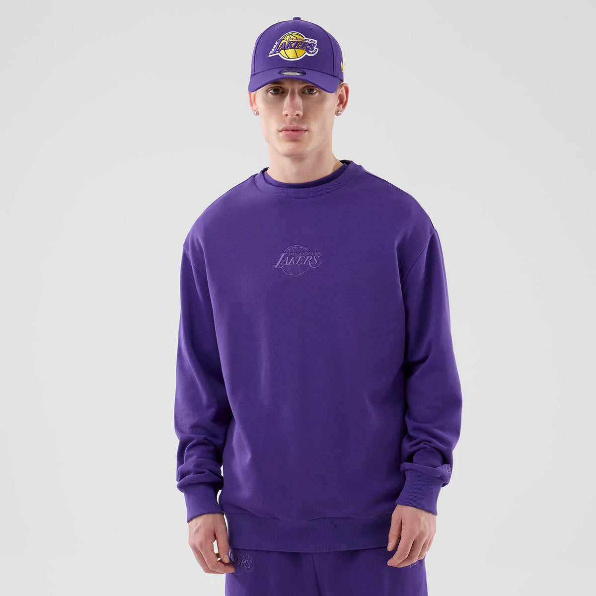 New Era LA Lakers NBA League Essential Purple Oversized Crew Neck Sweater, front view on a model paired with a purple Lakers cap, featuring embroidered Lakers logo on the chest and relaxed fit with ribbed cuffs and hem.
