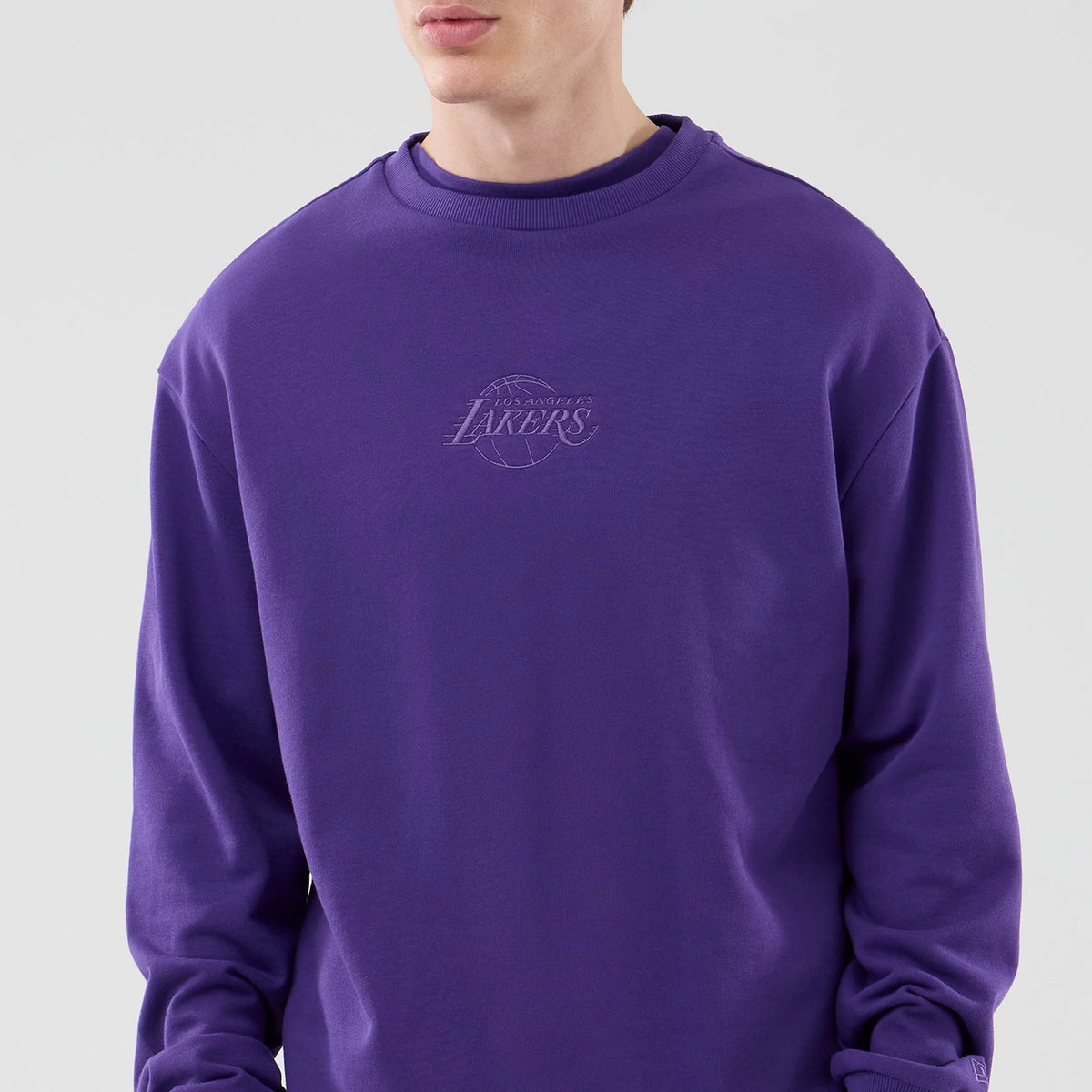 New Era LA Lakers NBA League Essential Purple Oversized Crew Neck Sweater, front view on a model showing the embroidered Lakers logo centred on the chest, with ribbed sleeves and neckline in a deep purple shade.