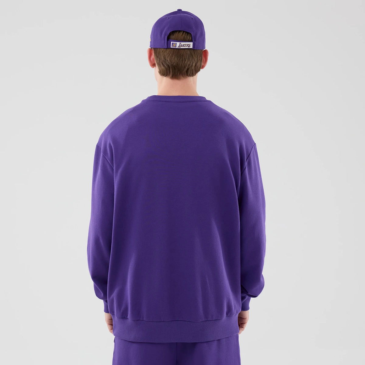 New Era LA Lakers NBA League Essential Purple Oversized Crew Neck Sweater, back view on a model with a matching purple cap, featuring a relaxed fit, a ribbed hem and cuffs, and an embroidered Lakers logo on the cap's back strap.