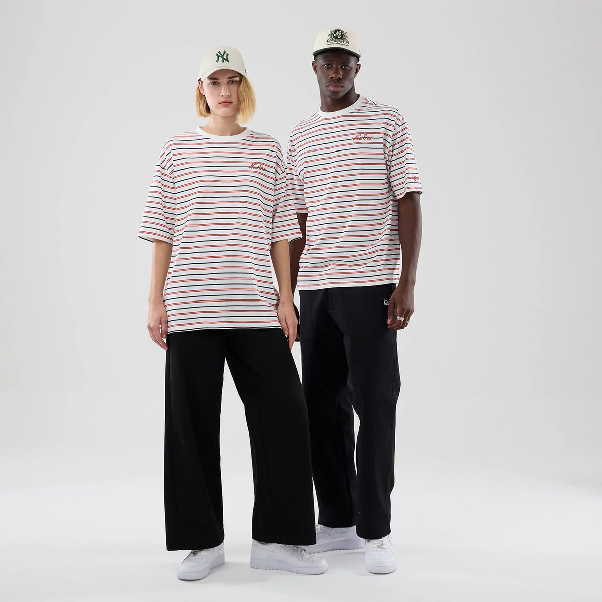 New Era Stripe White Oversized T-Shirt, front view on models featuring horizontal red and blue stripes, a red embroidered New Era logo on the chest left side, paired with black trousers and white trainers.