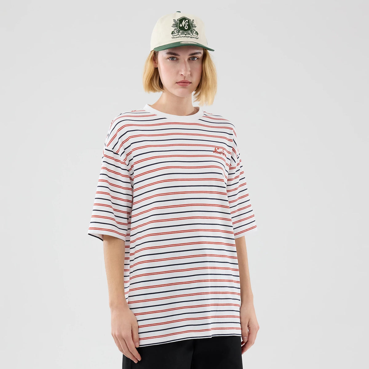 New Era Stripe White Oversized T-Shirt, front view on a model wearing a pale cap, featuring horizontal red and black stripes across the white fabric and a small embroidered New Era logo on the left chest.