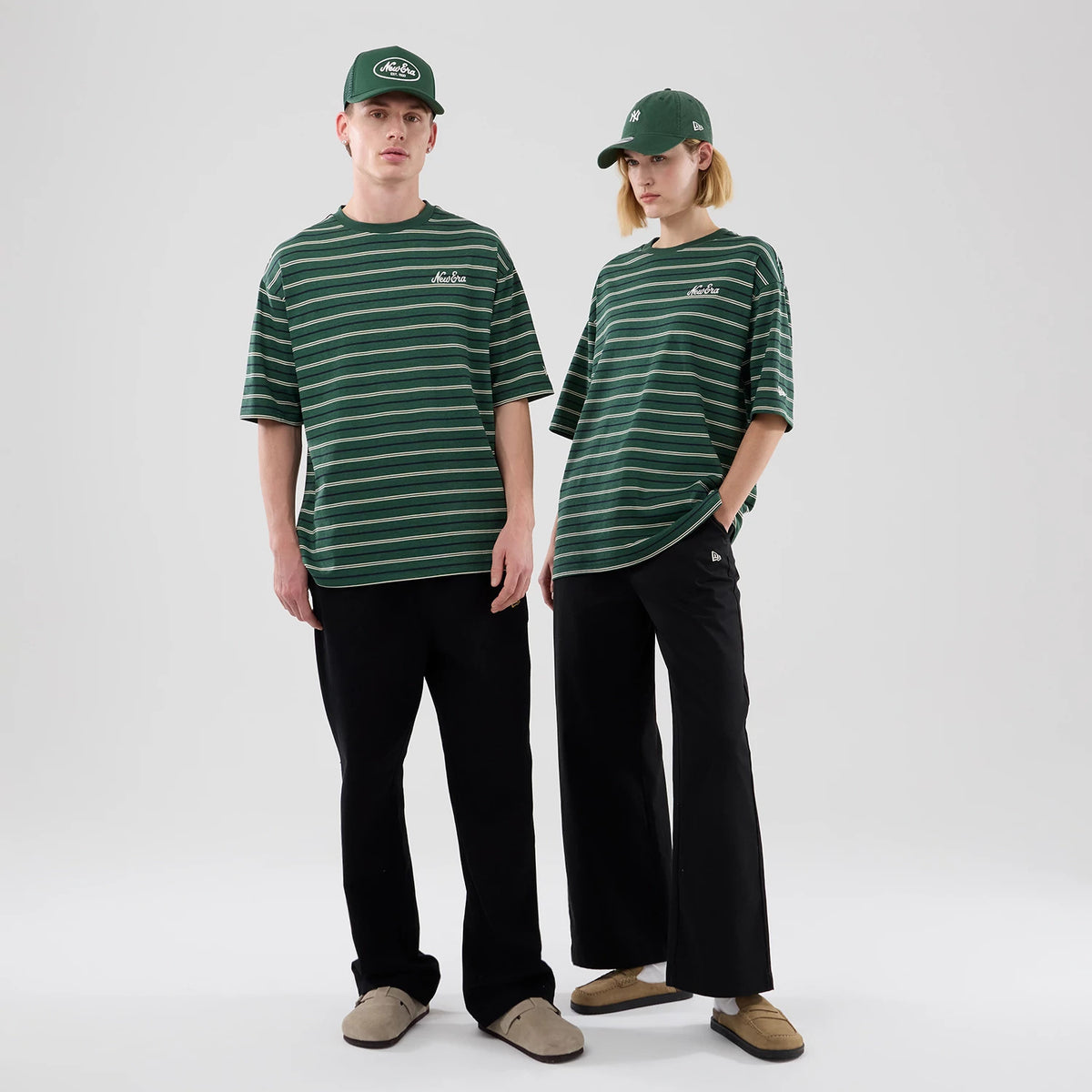 New Era Stripe Dark Green Oversized T-Shirt, on-model front view displaying alternating green and white horizontal stripes, a white embroidered New Era logo on the chest, and styled with black trousers and matching dark green New Era caps.