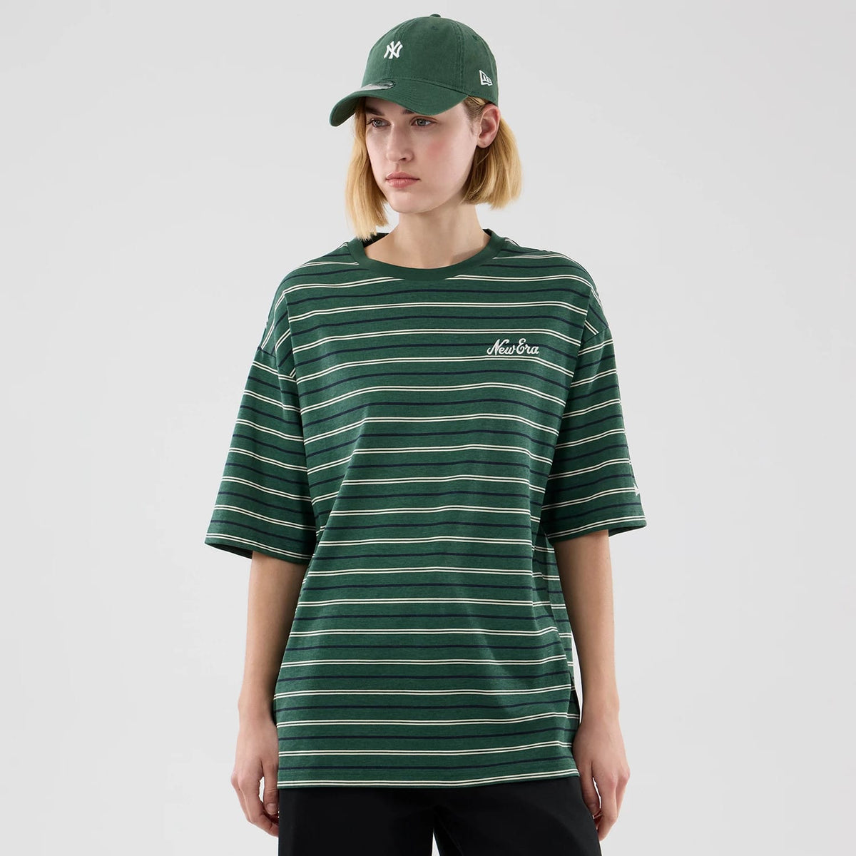 New Era Stripe Dark Green Oversized T-Shirt, front view on a model wearing a green cap, featuring horizontal white and yellow stripes across the dark green fabric, with the embroidered New Era logo in white on the left chest.
