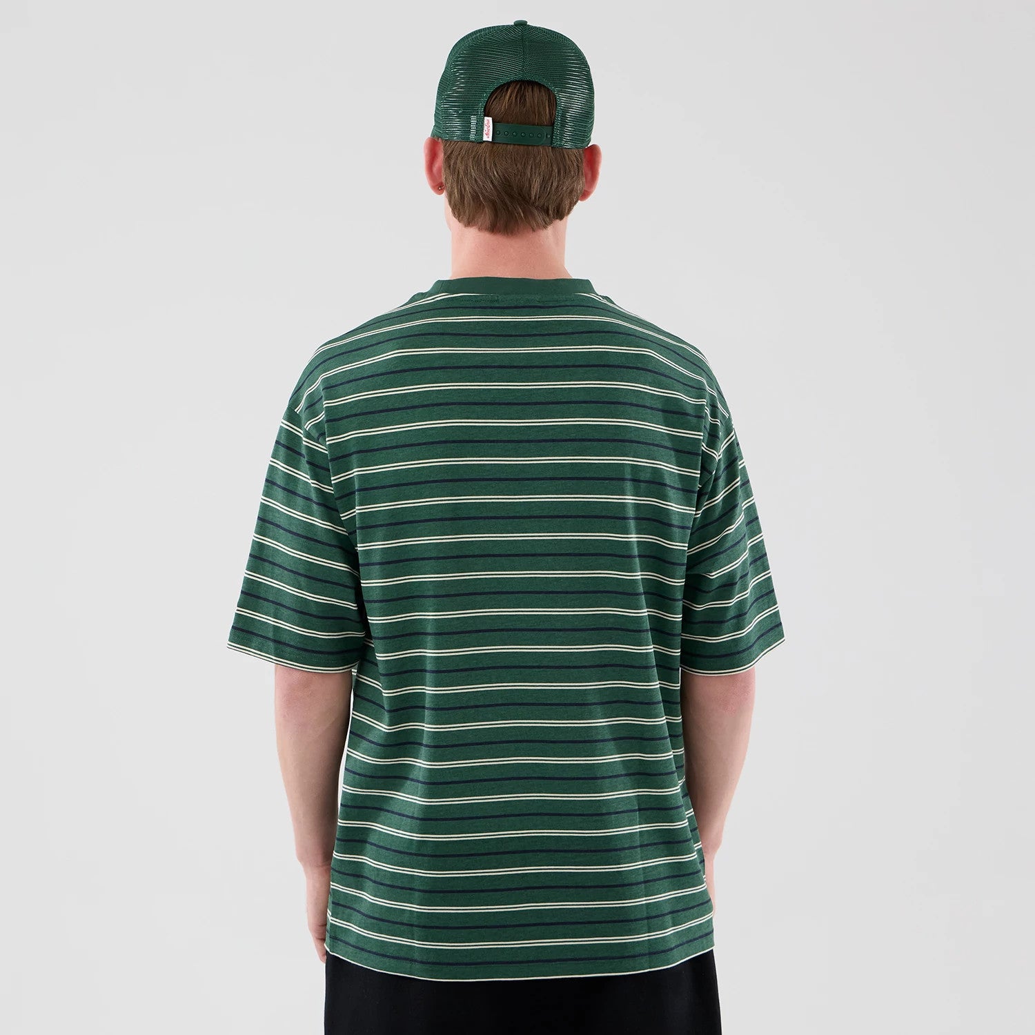 New Era Stripe Dark Green Oversized T-Shirt, back view on a model also wearing a matching cap, displaying horizontal green and white stripes across the back and sleeves, with a relaxed fit that falls loosely over the shoulders.