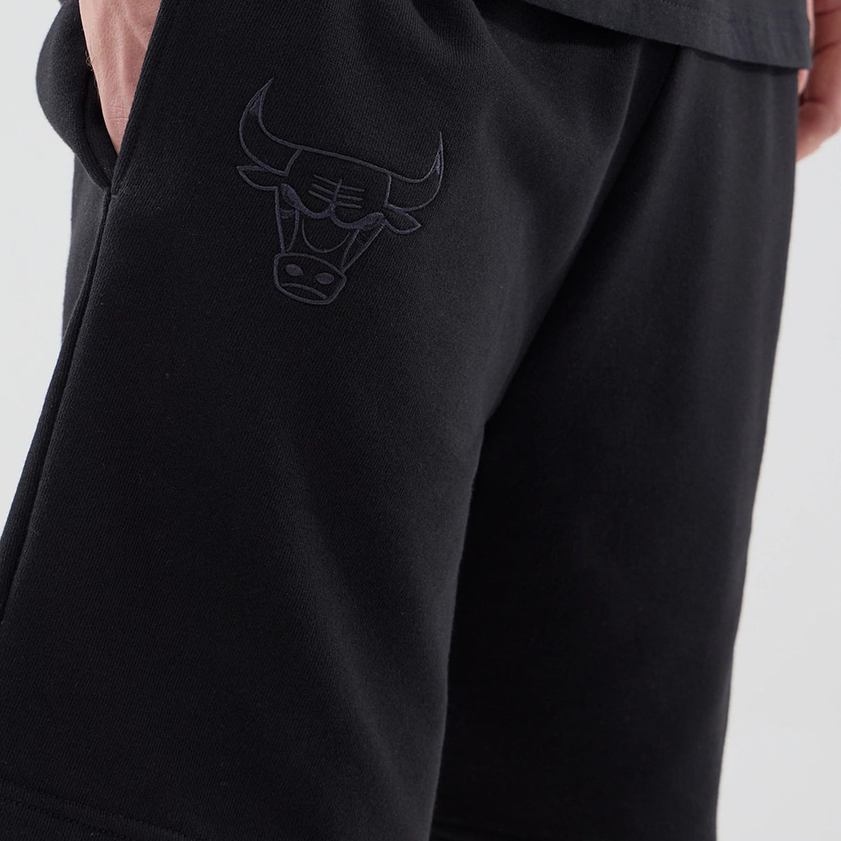 New Era Chicago Bulls NBA League Essential Black Oversized Shorts, front view showing the black embroidered Chicago Bulls logo on the left thigh, a loose-fitting silhouette, and a hand resting casually in the side pocket.