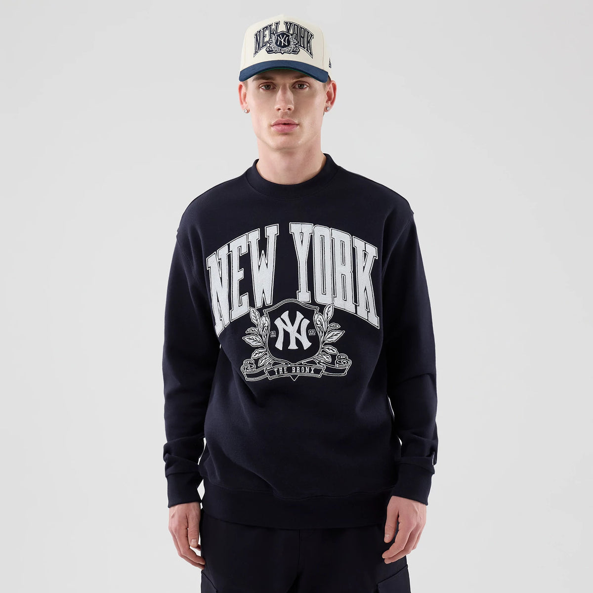 New Era New York Yankees MLB Arch Graphic Navy Oversized Crew Neck Sweater, front view on a model with a matching cap, featuring large white New York text and a central Yankees logo with laurel details on the chest.