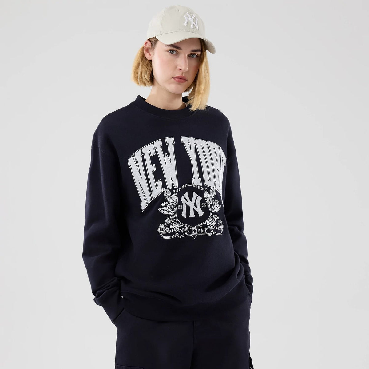 New Era New York Yankees MLB Arch Graphic Navy Oversized Crew Neck Sweater, front view on a model wearing a beige cap, featuring white New York lettering across the chest, the Yankees logo encircled by a decorative graphic, and long sleeves.