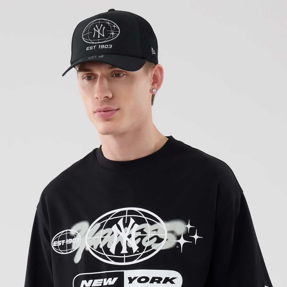 New Era New York Yankees MLB Logo Graphic Black Oversized T-Shirt, front view on a model featuring a prominent white graphic with a Yankees insignia on the chest, EST 1903 printed nearby, and a matching black cap with a raised logo.