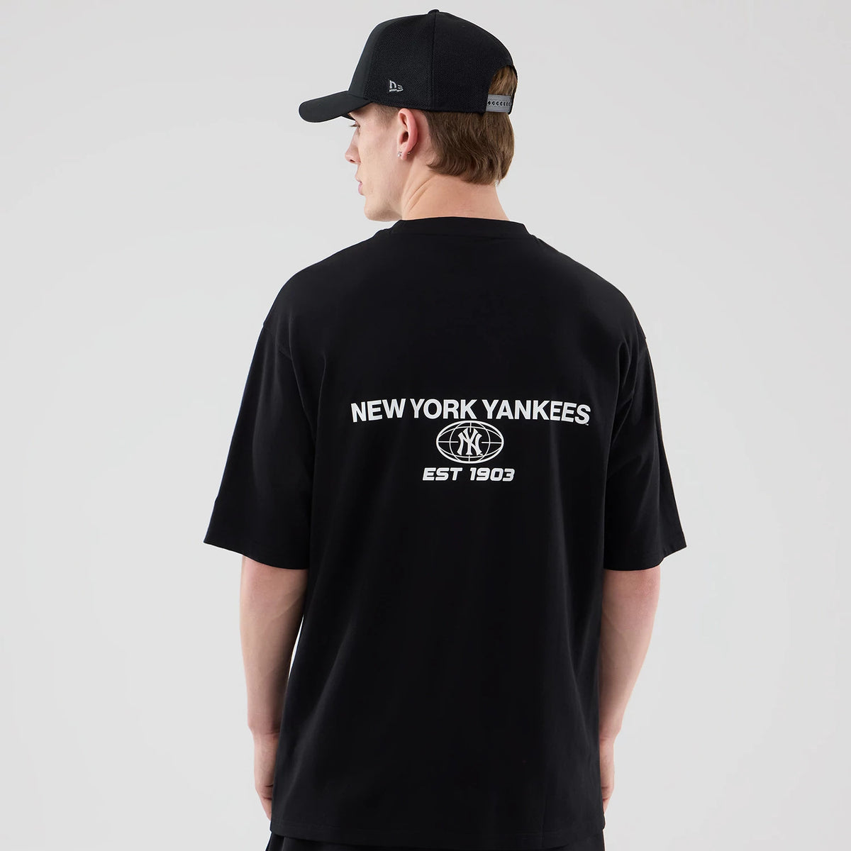 New Era New York Yankees MLB Logo Graphic Black Oversized T-Shirt, back view showing the white printed NEW YORK YANKEES text with the team logo and EST 1903 below, worn by a model styled with a matching black New Era cap.