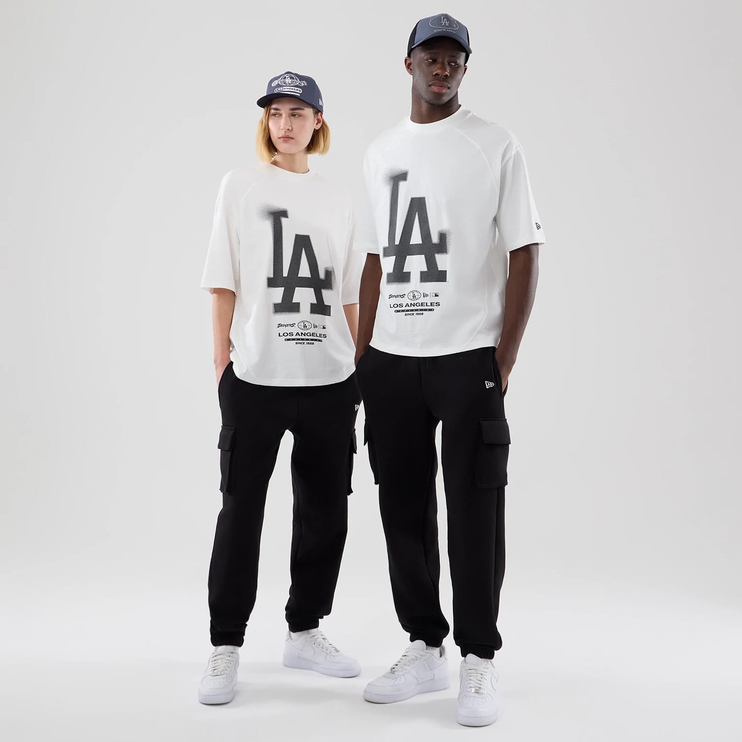 New Era LA Dodgers MLB Graphic White Boxy T-Shirt, front view on-model showing the large black LA logo spanning the chest, smaller team graphics below, and styled with black trousers and white trainers, both models also wearing matching caps.