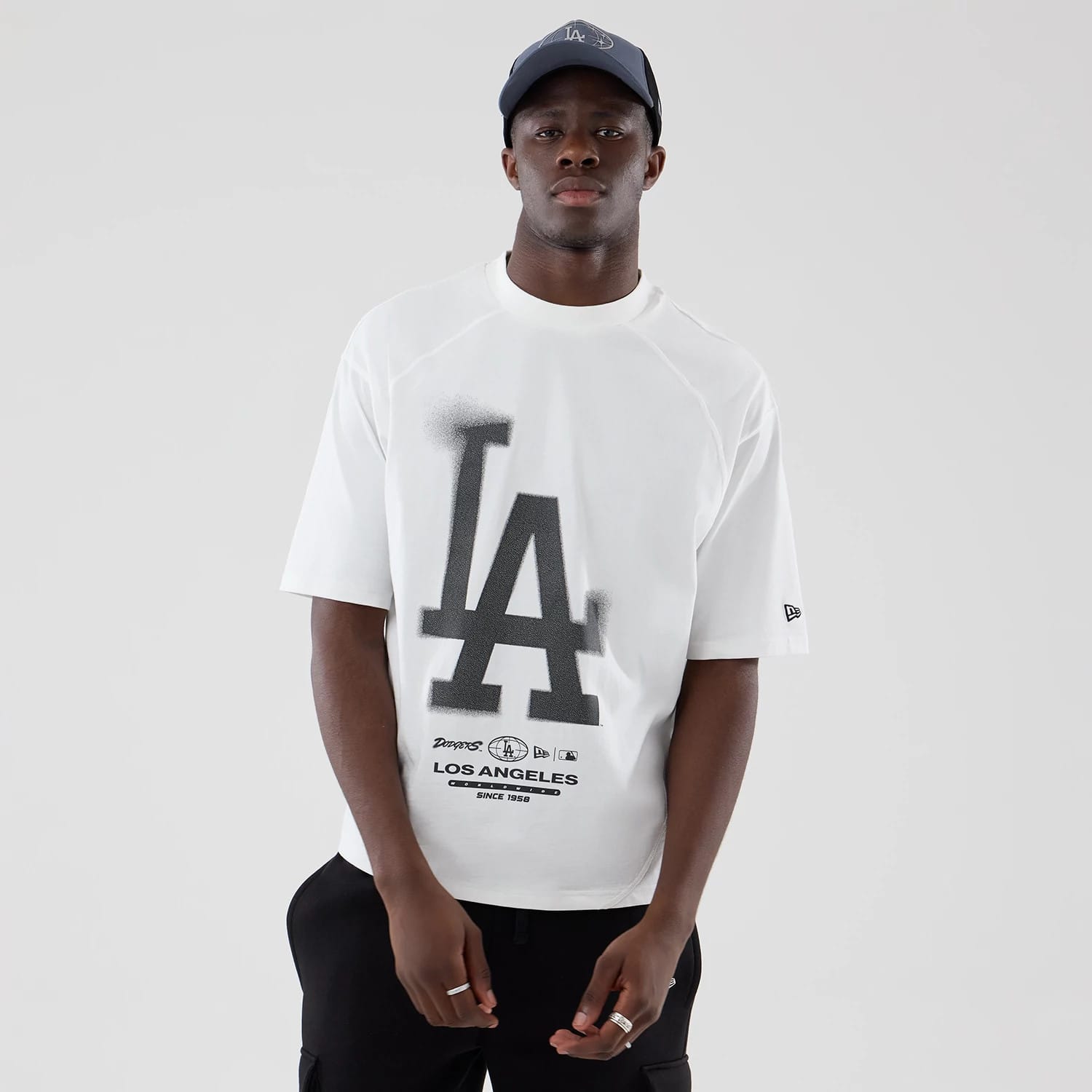 New Era LA Dodgers MLB Graphic White Boxy T-Shirt, front view on a model with a matching dark LA cap, featuring a large black LA logo on the chest, printed Dodgers graphics below and an embroidered New Era logo on the left sleeve.