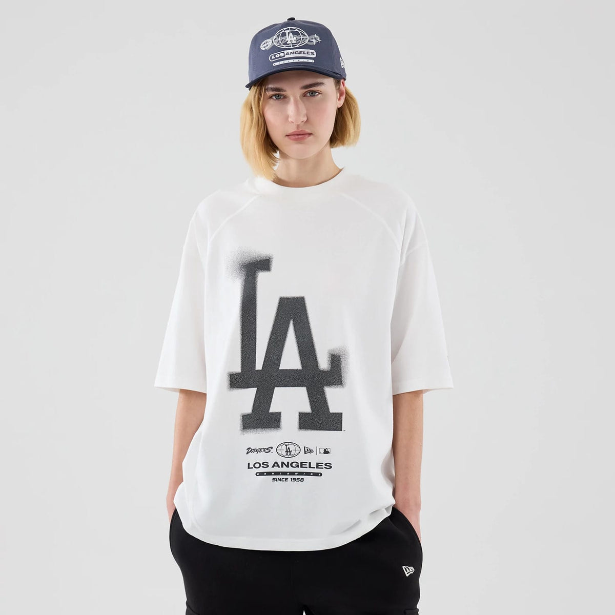 New Era LA Dodgers MLB Graphic White Boxy T-Shirt, front view on a model styled with a blue cap, featuring a large LA logo in black across the chest and additional team insignia graphics along the lower front of the white oversized tee.