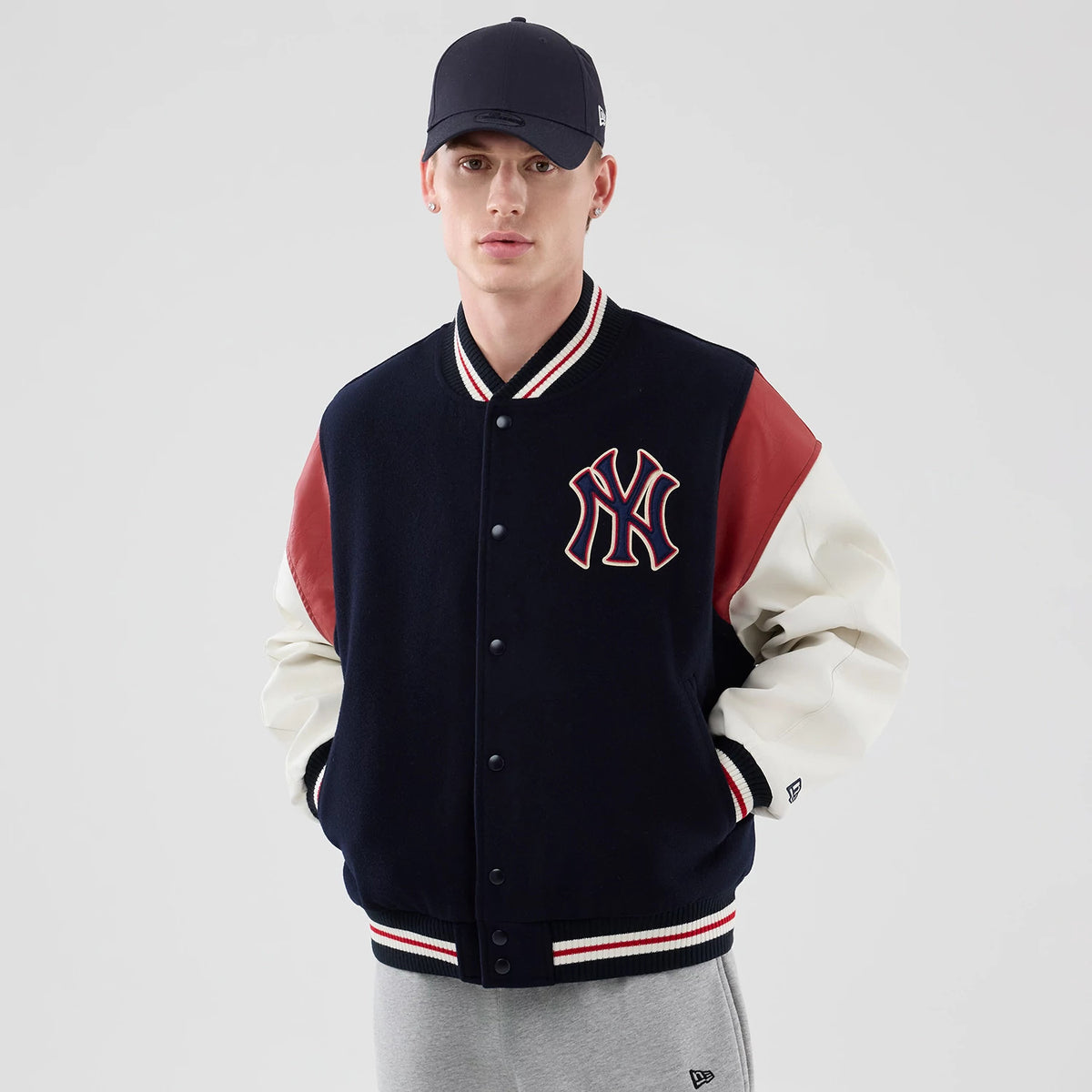 New Era New York Yankees MLB Varsity Navy Jacket, front view on a model styled with a matching cap, featuring a large embroidered NY logo on the left chest, red and white striped ribbed cuffs, and contrasting red and white sleeves.
