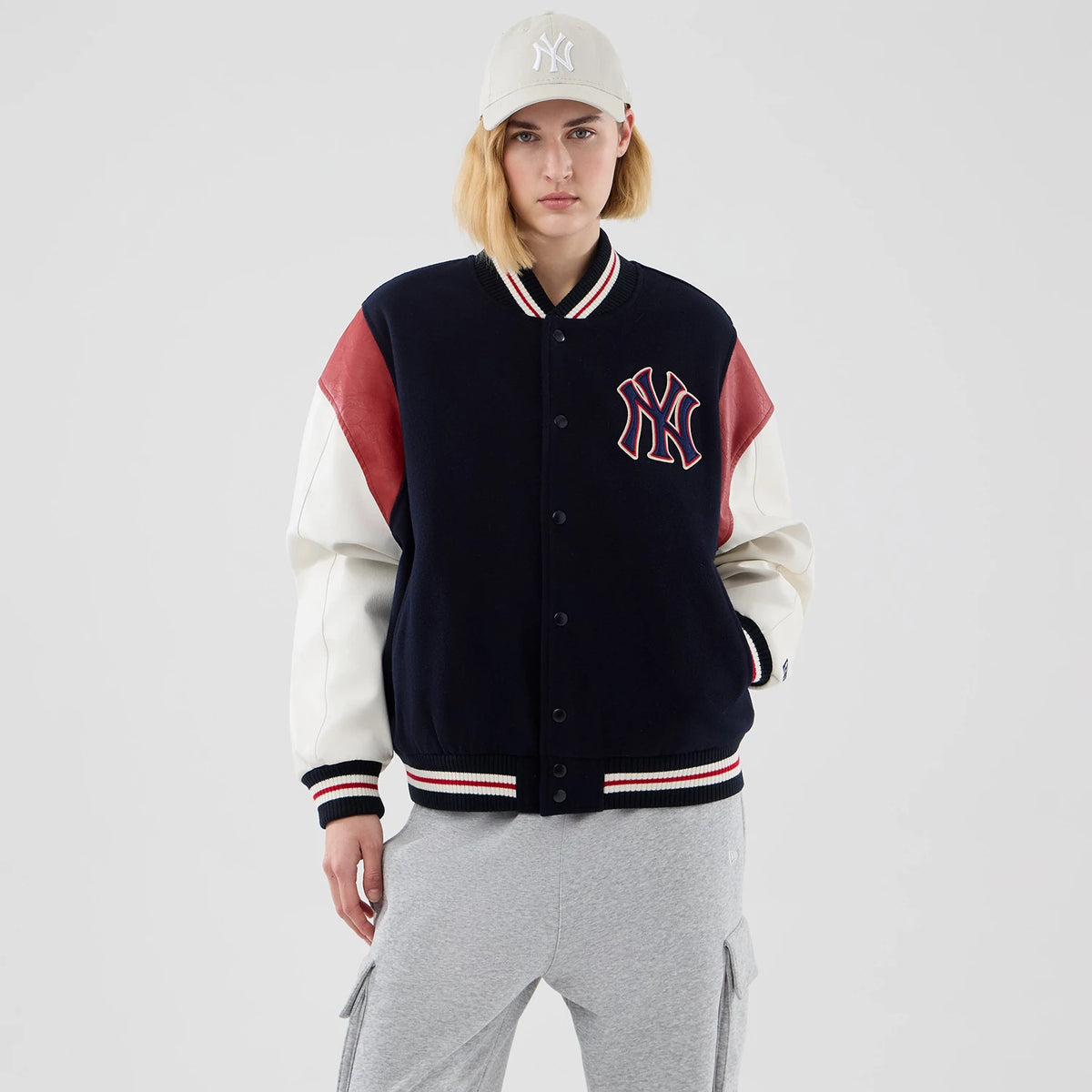 New Era New York Yankees MLB Varsity Navy Jacket, front view on-model showing a navy body with red and white striped trim, large embroidered NY logo on the left chest, red panels on the shoulders and white sleeves, styled with grey trousers.