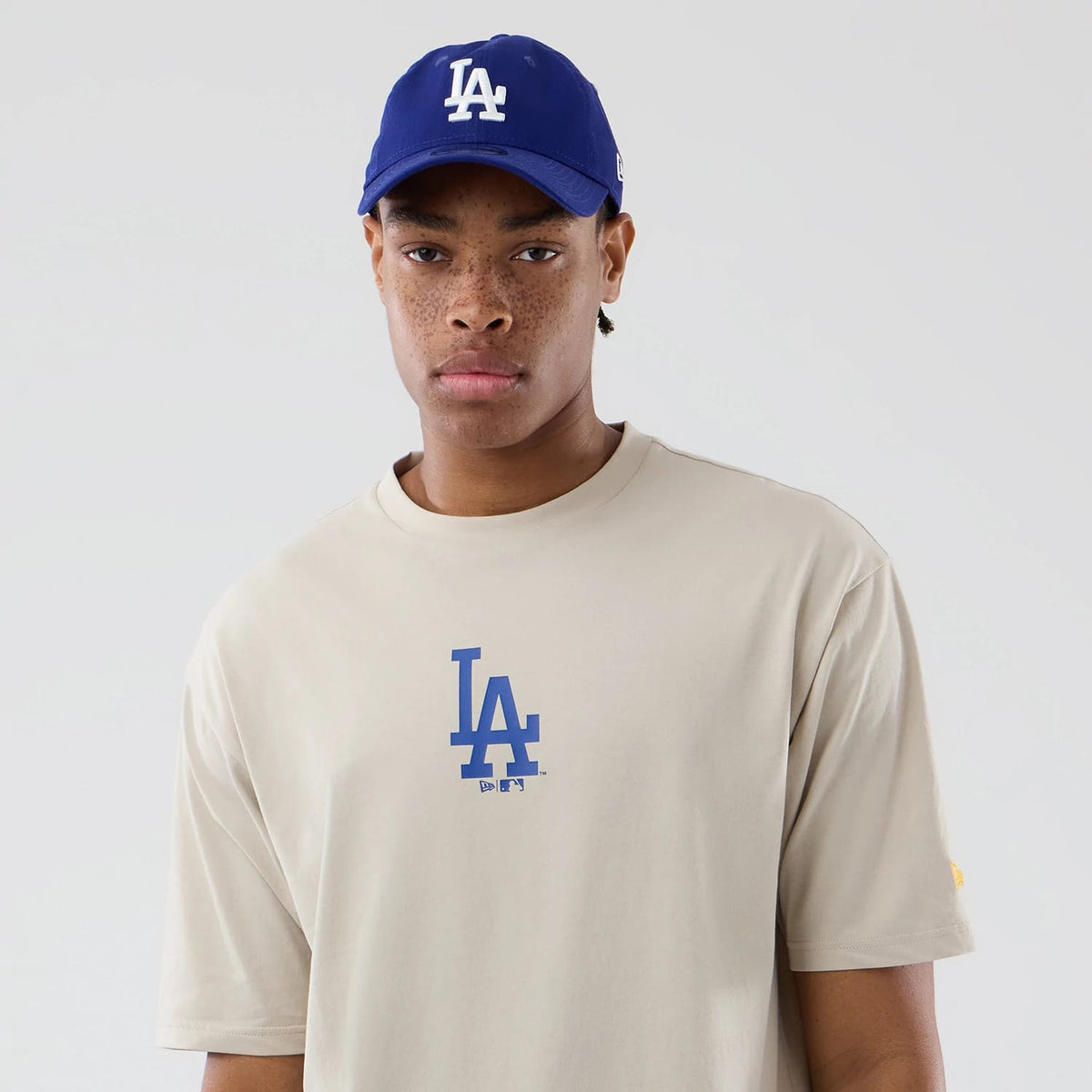 New Era LA Dodgers MLB Food Graphic Cream Oversized T-Shirt, front view on a model styled with a matching blue cap, featuring a blue printed LA Dodgers logo centred on the chest and a small yellow embroidered logo on the left sleeve.