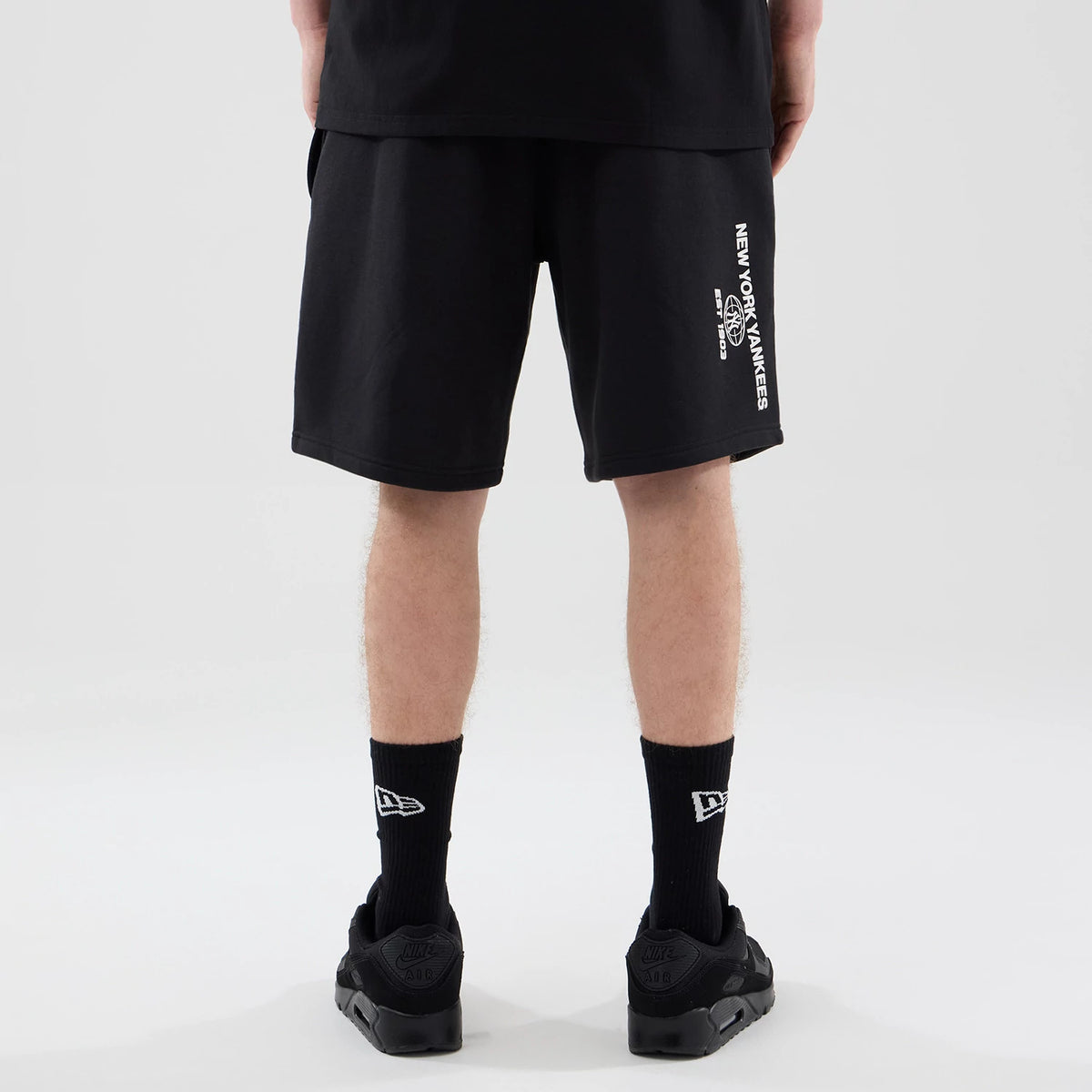 New Era New York Yankees MLB Logo Graphic Black Oversized Shorts, back view on a model featuring white printed New York Yankees lettering running vertically down the side, loose fit extending to the knees.