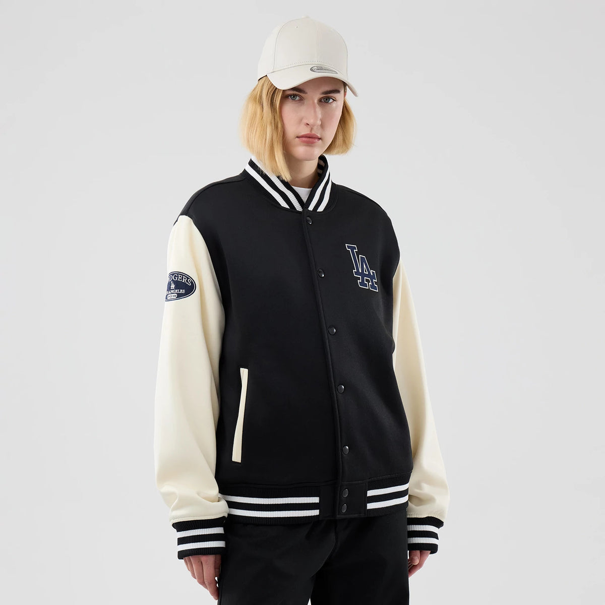 New Era LA Dodgers MLB Jersey Bomber Black Jacket, three-quarter view on a model showing the black body with contrast cream sleeves, an embroidered LA logo on the chest, and a Dodgers patch on the sleeve above the striped cuffs and collar.