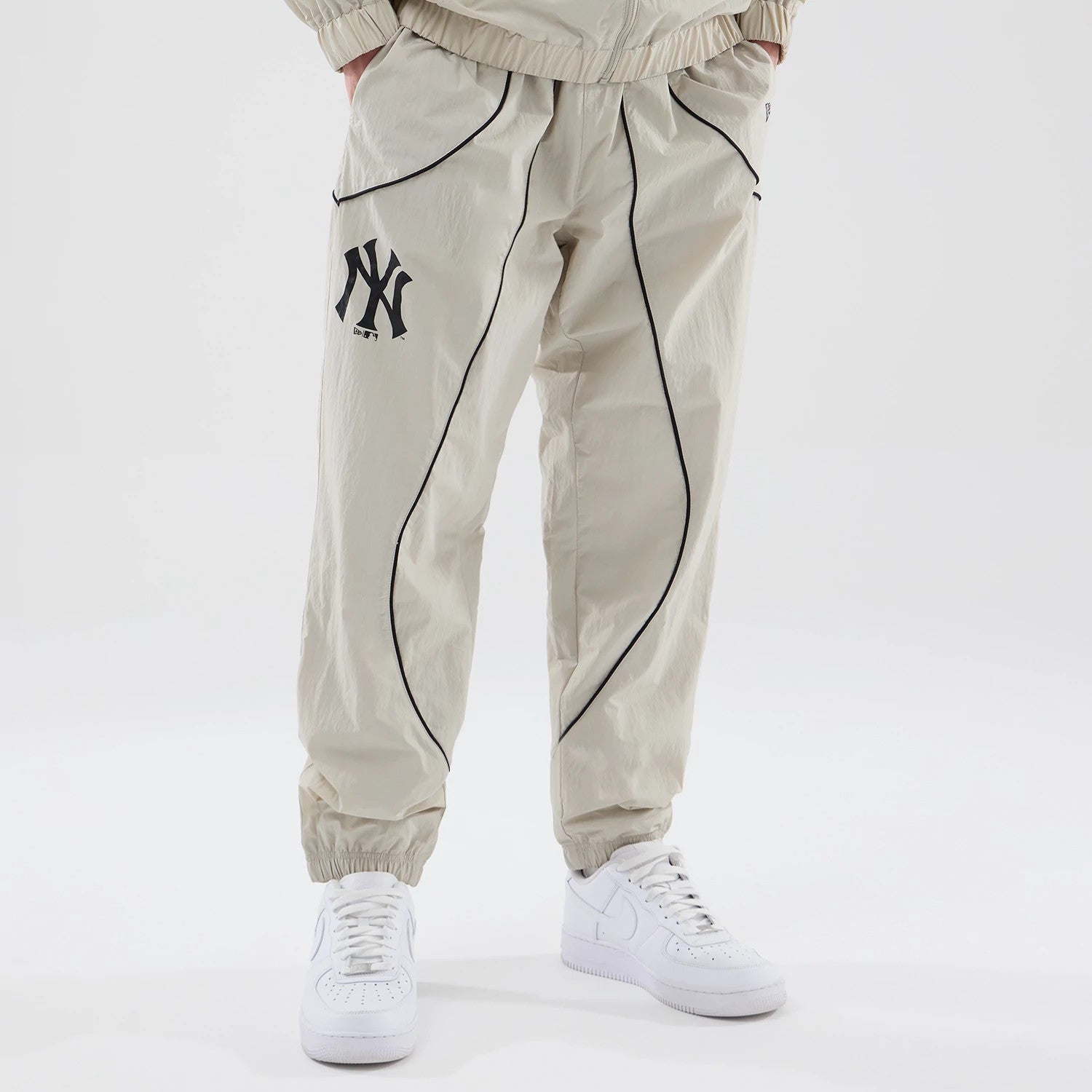 New Era New York Yankees MLB Piping Track Cream Track Pants, front view on a model showing black piping along the sides and front seams, a black Yankees logo on the left thigh, and elasticated cuffs above white trainers.